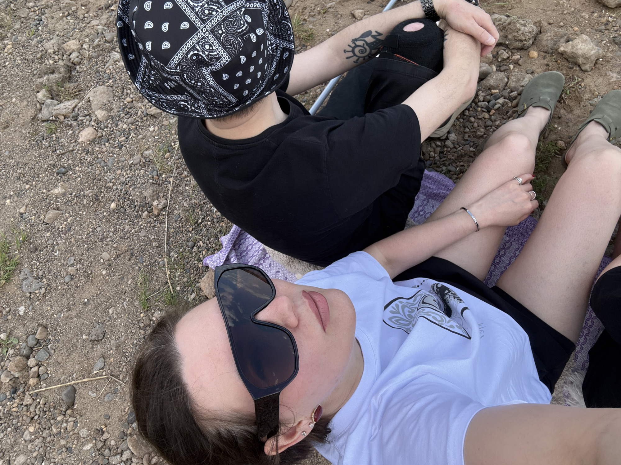 The image shows two people sitting on a purple blanket spread out on a pebbled surface, possibly a beach or riverbank. One person is wearing a white t-shirt, black shorts, and sunglasses. The other person is wearing a black t-shirt, black ripped jeans, a bandana bucket hat, and green shoes. The person in black is sitting with crossed legs, and the person in white is lying back. The location is likely near Doksha, Russia, given that was where the image was taken.