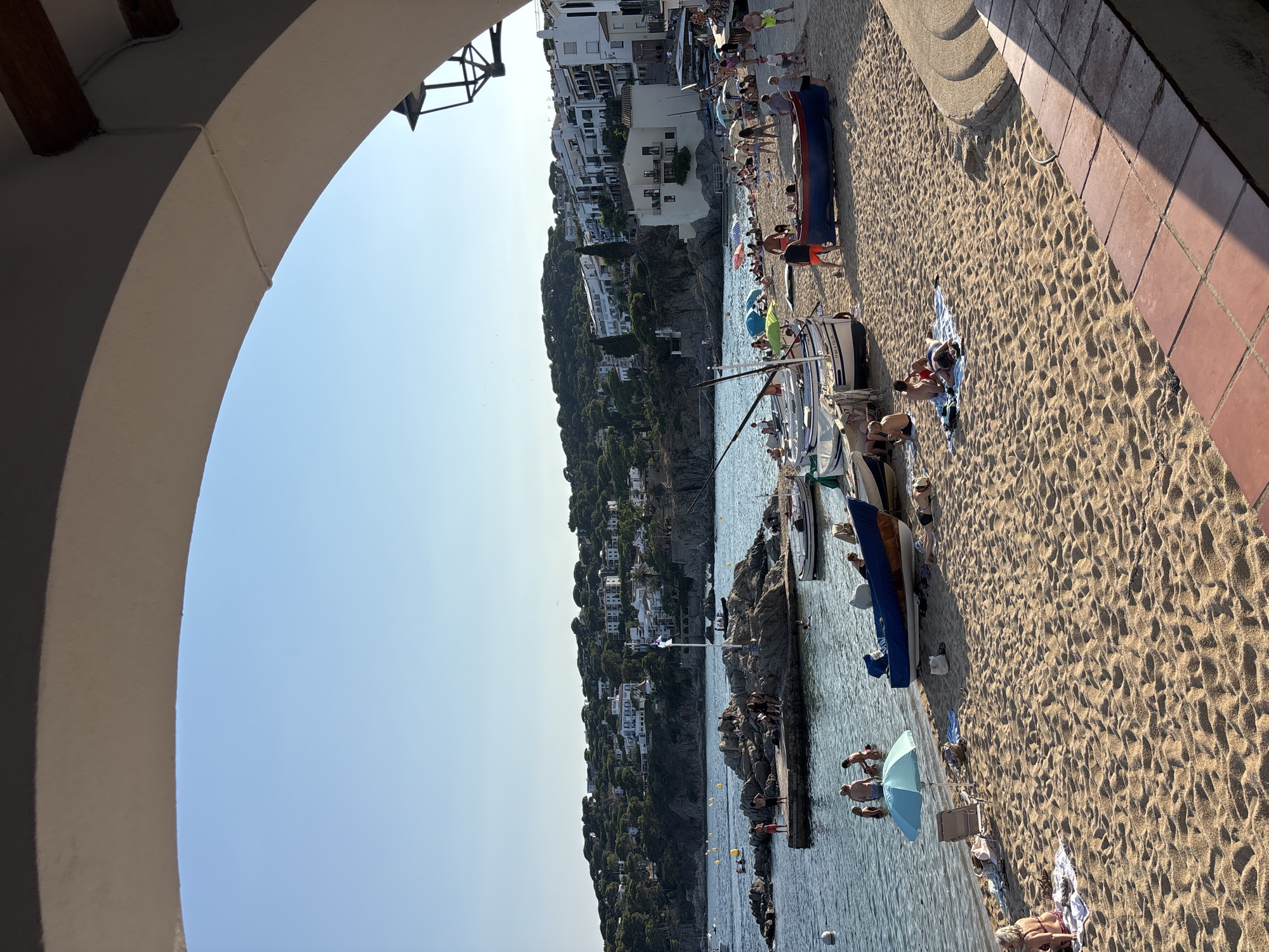 The image depicts a sunny day at a beach in Calella De Palafrugell, Spain, viewed through an architectural archway. The sandy beach is populated with numerous people engaged in various beach activities. Some individuals are sunbathing on towels and chairs, while others are in the shallow water, swimming or wading. Several colorful umbrellas provide shade.
A collection of boats, both small fishing vessels and larger pleasure boats, are either moored near the shore or pulled up onto the sand. One notable boat is a blue and red fishing boat resting on the beach. In the background, the clear blue sea stretches towards a rocky outcrop dotted with people. Beyond the outcrop, a picturesque coastal town with white buildings and lush green hills rises against the horizon. The sky is clear and bright blue, indicative of good weather. The foreground features a tiled walkway and part of the archway framing the scene.