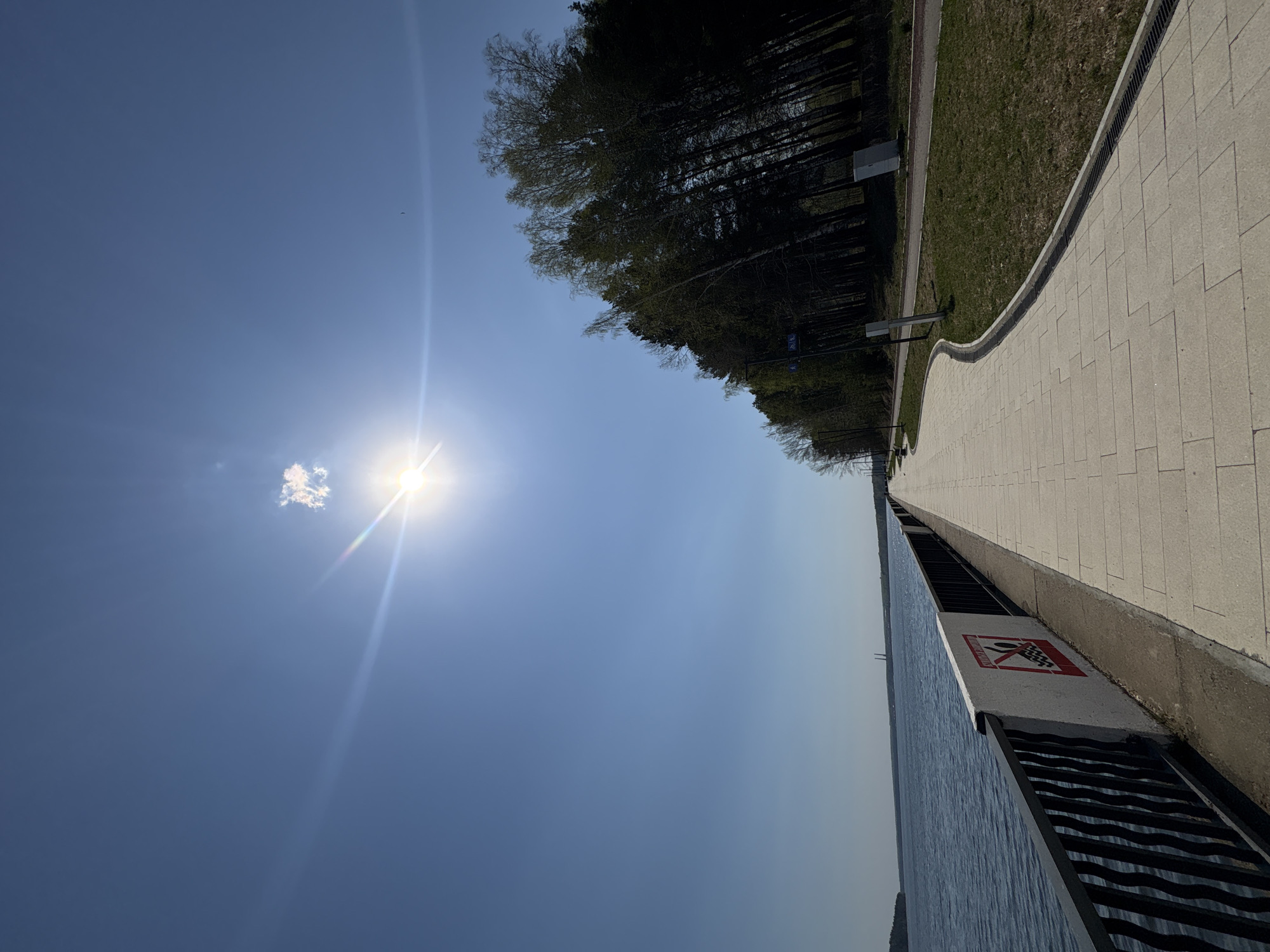 The image shows a daytime view of a waterfront area in Chaikovsky, Russia, with a blue sky and bright sun partially obscured by a small cloud. A paved walkway runs along the right side of the image, bordering a grassy area and trees beyond. A metal railing separates the walkway from the water. A sign with a red and white symbol is visible on a section of the wall near the railing. The water appears calm.