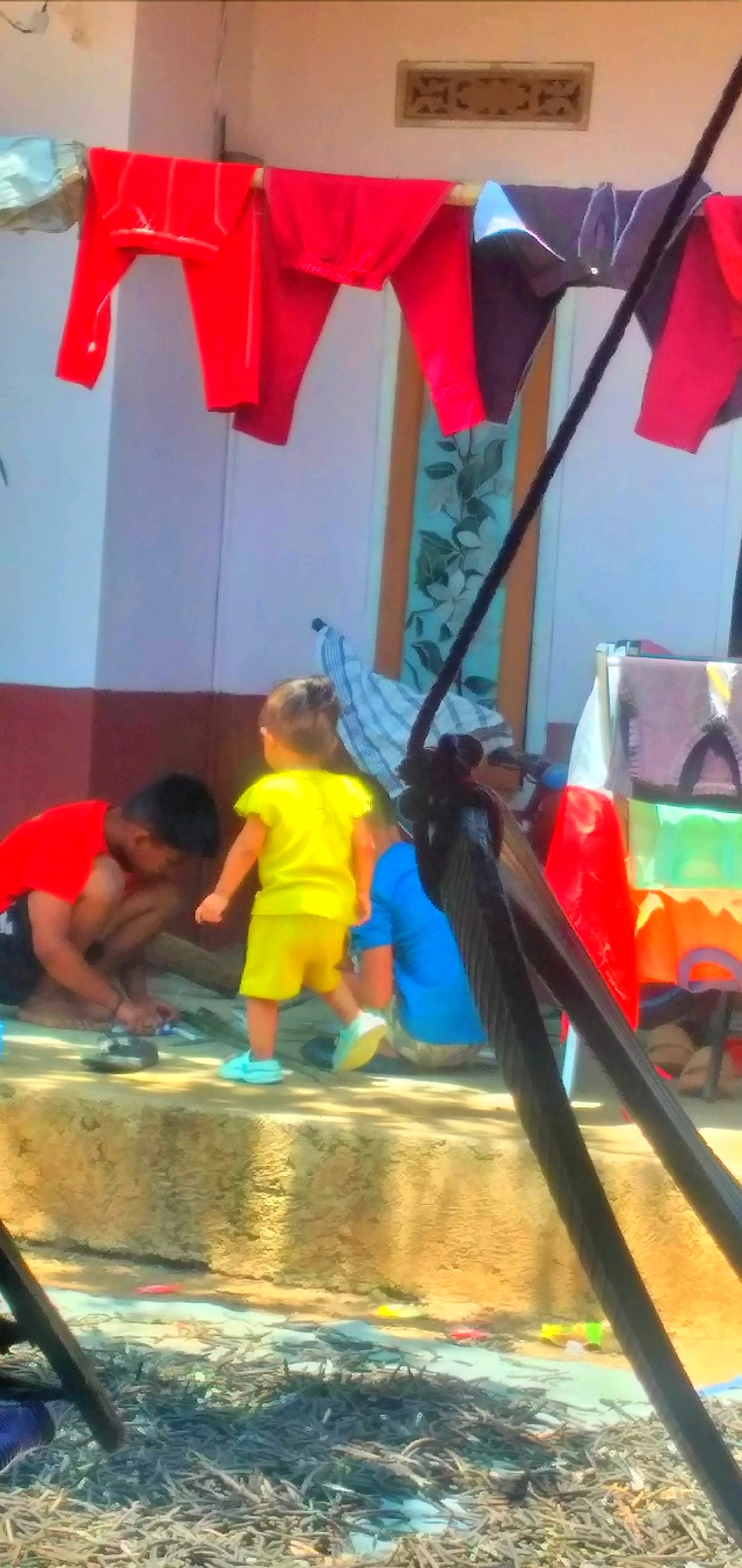A slightly blurred, medium shot captures three children outdoors in Rahong, Indonesia, near a building with clothes drying on a line overhead. In the foreground, a young boy wearing a red shirt and dark shorts is crouched down, appearing to play with something on the ground. To his right, a toddler in a bright yellow t-shirt and shorts stands facing away from the camera, also seemingly engaged with the ground. Further to the right and slightly behind the toddler, another child is visible, wearing a blue shirt and patterned shorts, also facing away. A clothesline is strung across the upper portion of the image, holding several red garments and some darker items. The ground surface is uneven and appears to be paved or a concrete step. The background includes a building wall and a doorway with a decorative panel.