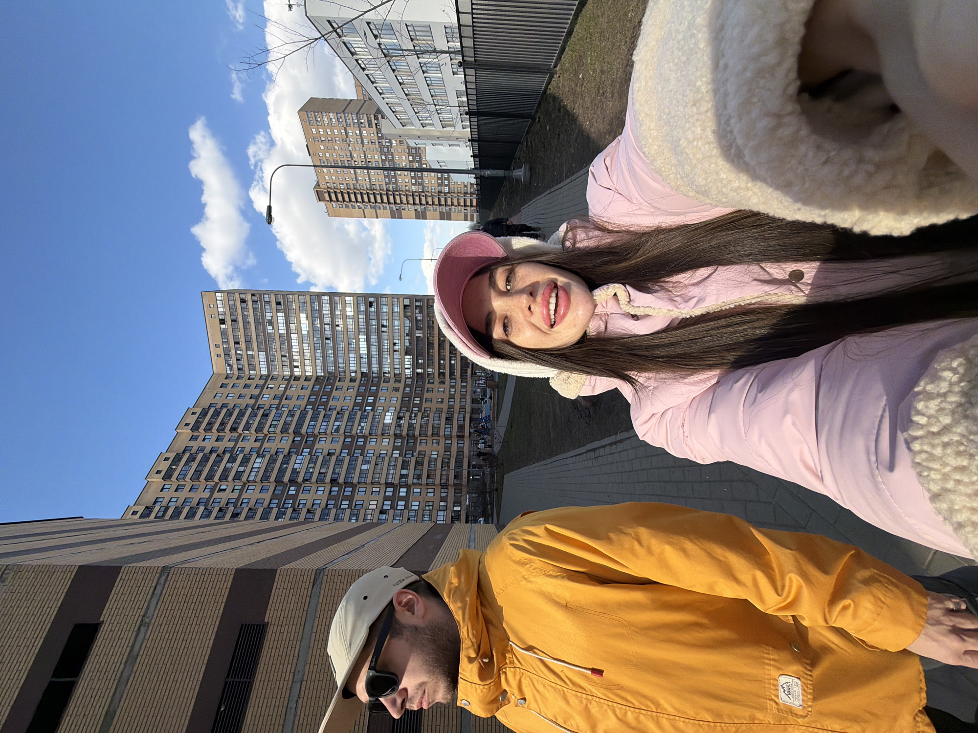 A selfie captures two individuals walking outdoors on a paved sidewalk in Saint Petersburg, Russia, under a clear blue sky with scattered white clouds.
In the foreground, a female with long dark hair, wearing a pink hooded jacket with a cream sherpa lining and a pink baseball cap beneath the hood, smiles directly at the camera while holding it with her extended right arm. Her lips are visibly enhanced and colored pink. To her left and slightly behind, a male subject is depicted in profile, looking towards the left, away from the camera. He wears a light-colored baseball cap, dark sunglasses, and a bright yellow hooded jacket with a white drawstring. A white "Vans" brand patch is visible on his left chest pocket. A ring is visible on the ring finger of his left hand.
The environment features a series of modern, multi-story residential buildings. A large, brown brick-patterned building with numerous windows dominates the background, curving to the right. To the far left, another tall, light-brown building with horizontal darker stripes is partially visible. Further down the street, a taller, slender brown building stands, and a grey/white multi-story building with a black fence in front of it is visible on the right side of the frame. A black street lamp post with a single light fixture is positioned in the mid-ground. The ground alongside the sidewalk consists of dry grass and bare dirt, with a few bare tree branches indicating a late winter or early spring season. Distant figures can be seen walking on the sidewalk further into the background.