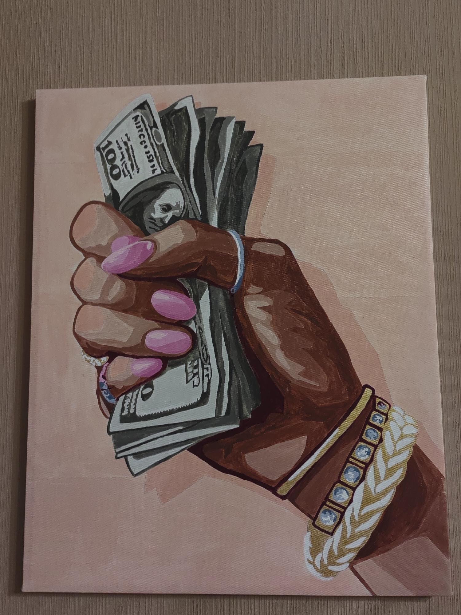 An oil painting on canvas depicts a hand holding a fan of US hundred-dollar bills. The hand is dark-skinned with long, pink, almond-shaped fingernails. Several rings are visible on the fingers, and a blue band is worn on the wrist. A thick bracelet with gold accents and clear stones adorns the forearm. The background is a pale pink, and the painting is framed by a wooden surface. The image does not provide any information about the city of Ostafievo, Russia, or any specific location within it.