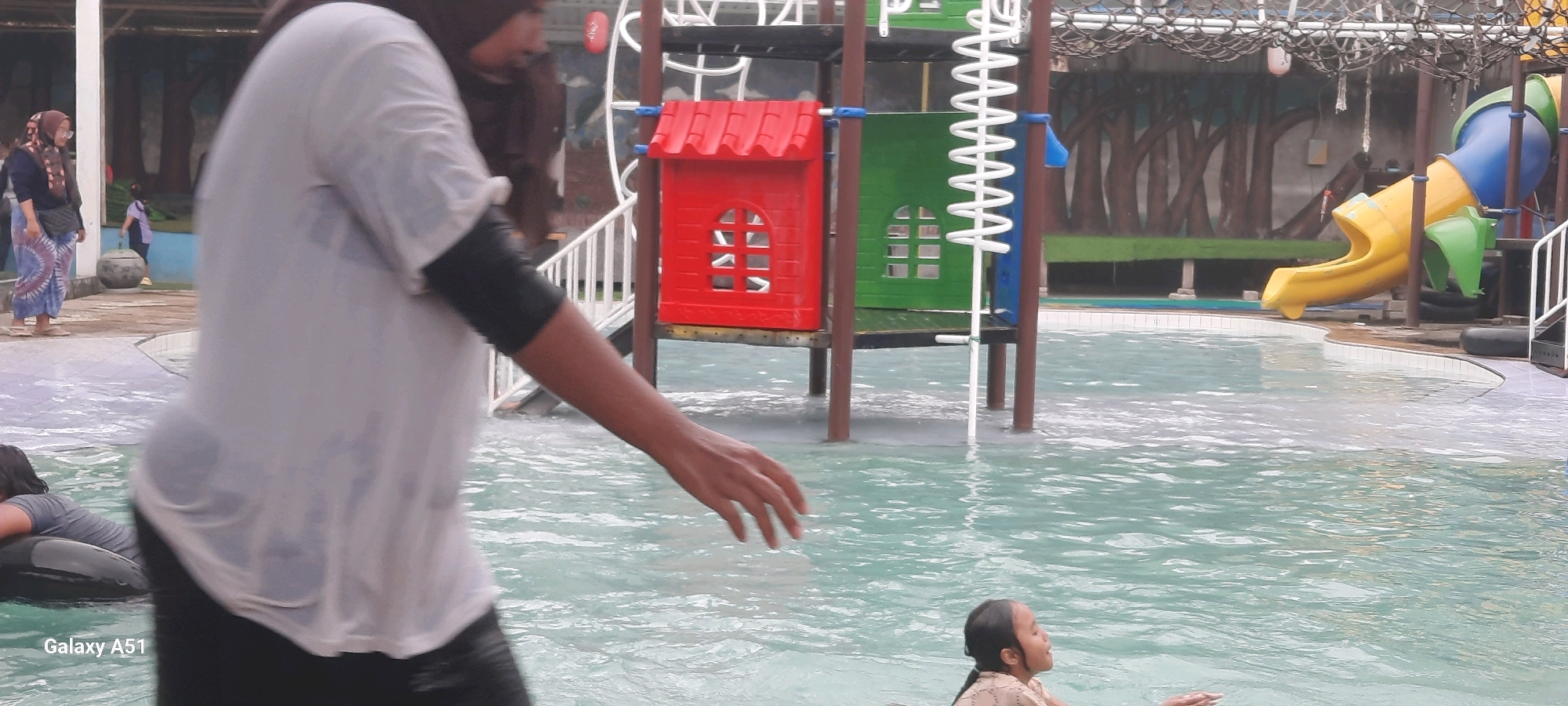The image depicts a scene at a water park, likely in Popoh, Indonesia, given the provided context. In the foreground, a person wearing a white t-shirt and dark leggings is partially visible, their arm extended into the shallow water. To the left, another individual is seen from the chest up, lying on an inflatable tube. In the mid-ground, a young girl with dark hair is partially submerged in the water, looking upwards with her eyes closed. Behind her, a colorful playground structure with red and green elements is visible, along with a yellow and blue water slide. In the background, a woman wearing a hijab and a patterned dress is standing near a large, dark, round object, possibly a planter or a decorative element. The water appears to be a light turquoise color, and the surroundings suggest an outdoor recreational area with trees depicted in the background mural. The overall impression is a lively, family-oriented environment at a water park.