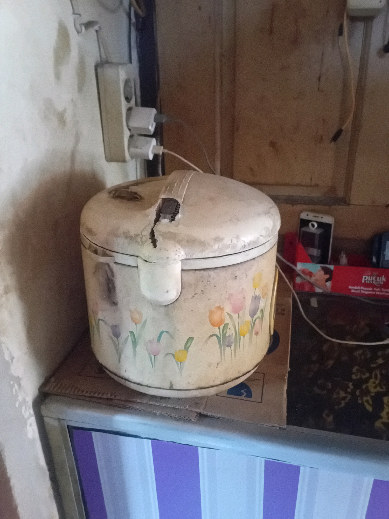 A vintage rice cooker, adorned with a floral pattern of tulips in various colors, sits on a cardboard box. The rice cooker is positioned on a shelf or counter in what appears to be a kitchen or storage area. To the left of the rice cooker, a white electrical outlet strip is mounted on a textured, off-white wall. Two white power adapters are plugged into the outlet strip. To the right of the rice cooker, a smartphone is visible next to a red box with text that reads "Pucuk Harum." The background consists of a wooden surface, likely a cabinet or wall panel, with visible grain and some smudges. The overall environment seems modest, possibly a home or small establishment in Pekanbaru, Indonesia. The lighting is dim, casting shadows and highlighting the worn surfaces of the objects.