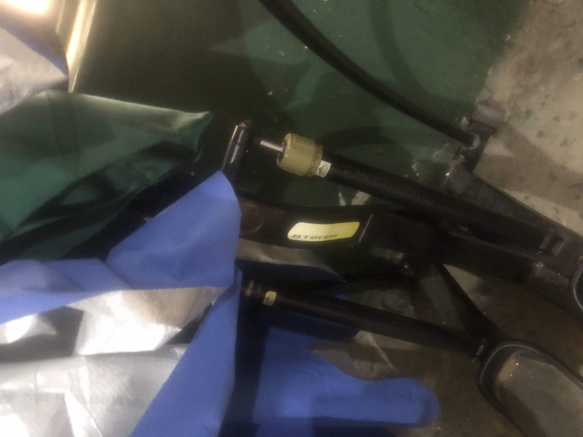 The image shows a close-up view of a piece of exercise equipment, likely a stationary bike or elliptical, partially covered by blue and green fabric. The equipment is black and features metallic components, including pedals and an adjustable cylinder. A yellow sticker with the text "ST2000" is visible on the equipment. The background is dimly lit and appears to be a wall painted green, with a concrete floor that has some debris or water stains. The overall scene suggests the equipment is stored in a non-ideal environment, possibly a garage or storage area. There are no people visible in the image, and no specific Jalingo landmarks are identifiable.