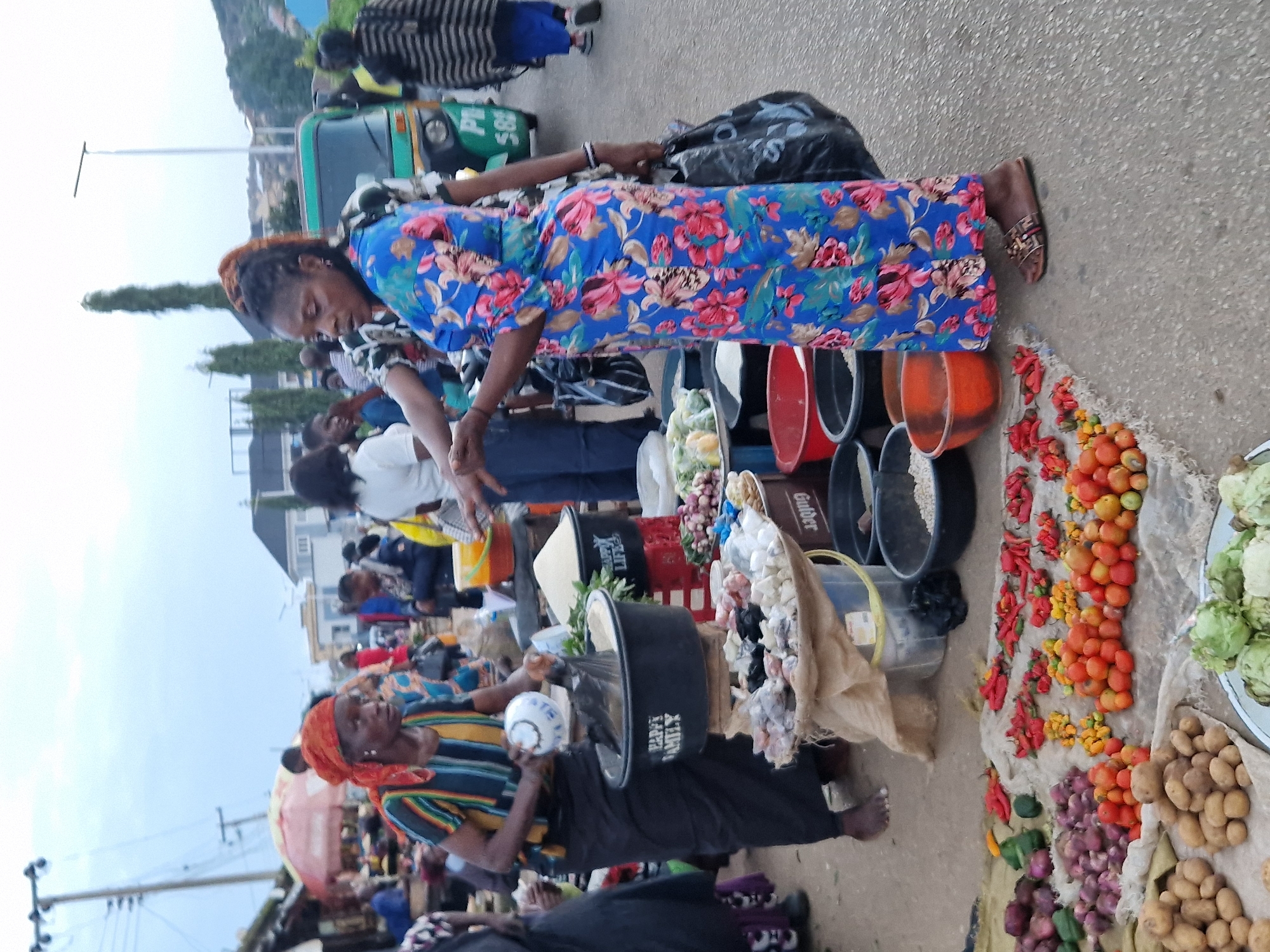 The image depicts an outdoor market scene in Kabong, Nigeria. In the foreground, a variety of fresh produce, including tomatoes, peppers, onions, and potatoes, are displayed on a cloth laid out on the ground. Several people are present in the market, engaged in transactions or moving through the space.
One woman, wearing a blue floral dress and sandals, is standing and holding a black plastic bag. She appears to be interacting with another woman who is partially visible. The second woman, in the left of the frame, wears a striped top and an orange headscarf, and is holding a white bowl over a large black basin. This basin contains a white granular substance, possibly rice or flour, and has "HAPPY FAMILY" inscribed on it.
Behind these two women, other market stalls and people are visible. There are various containers, including buckets and crates, holding food items and goods. A green motorized tricycle, commonly known as a Keke Napep, is parked nearby, suggesting a mode of transportation or a vendor's setup. The background shows some buildings, trees, and a cloudy sky. The ground surface is asphalt or concrete, indicative of a paved market area. The overall atmosphere suggests a bustling marketplace.