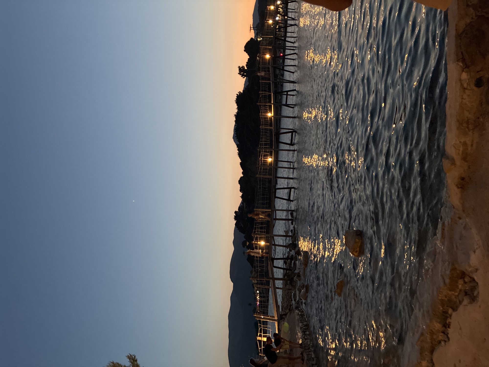 The image depicts a waterfront scene at dusk. A wooden pier extends over the water, supported by stilts. String lights illuminate the pier, casting reflections on the water's surface. Several people are visible on and near the pier, some appearing to walk or stand. In the background, silhouetted hills and trees are visible against a gradient sky that transitions from a pale blue at the top to an orange hue near the horizon. The water is slightly rippled, with rocks visible near the shore. The overall ambiance suggests a peaceful evening setting, possibly at a coastal town. The location is in the city of Laganas Municipal Unit, Greece.