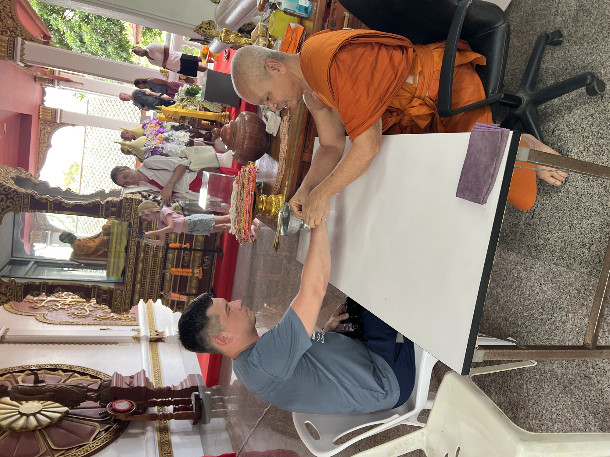 A Buddhist monk in orange robes sits across a white table from a man wearing a grey t-shirt. The monk is holding the man's left wrist, appearing to tie a string around it. The man is seated in a white plastic chair, while the monk is in a black office chair. The table has a silver bowl and a purple cloth on it. In the background, other people are visible, along with religious decorations and a statue of Buddha. The scene is indoors, likely within a temple or religious building in Baan Hua Thanon, Thailand.
