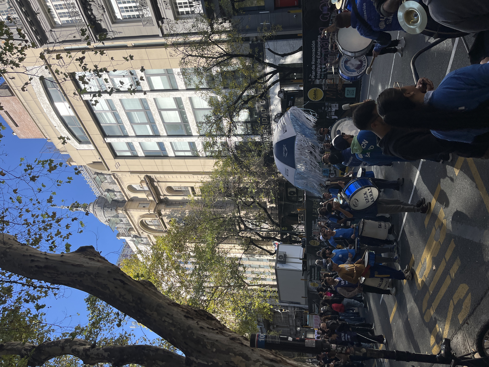 This media file captures an outdoor urban scene in Buenos Aires, Argentina, depicting a public gathering or demonstration under a clear blue sky. The image shows a street lined with buildings of varying architectural styles, from ornate, classical structures with domes and intricate detailing to more modern edifices with large windows. In the foreground, a prominent tree trunk and branches partially frame the view.
The main subjects are a large group of people participating in what appears to be a parade, protest, or marching band. Many individuals are dressed in blue shirts, some bearing the logos "U.P.C.N." and "SALUD". They are actively playing percussion instruments, including large drums with "U.P.C.N. A.N.M.A.T." and "SALUD" visible on their surfaces, and cymbals. A distinctive blue and white fringed umbrella is also visible among the crowd. The street features yellow road markings.
The event is taking place during the daytime, evidenced by the bright sunlight and distinct shadows. The weather is sunny and clear. Visible text on signs includes "Plan de Renovación Integral de Estaciones," "Subte" (referring to the Buenos Aires subway system), "UNIVERSIDAD," and brand names like "CLARO." Other texts such as "LAS PIBAS," "ORGANIZACIÓN," and "VENCE" are discernible on various banners or signs. The presence of "U.P.C.N." (a major Argentine trade union) and "A.N.M.A.T." suggests this could be a demonstration related to public sector employees, health services, or education in Argentina. The general atmosphere appears active and organized.