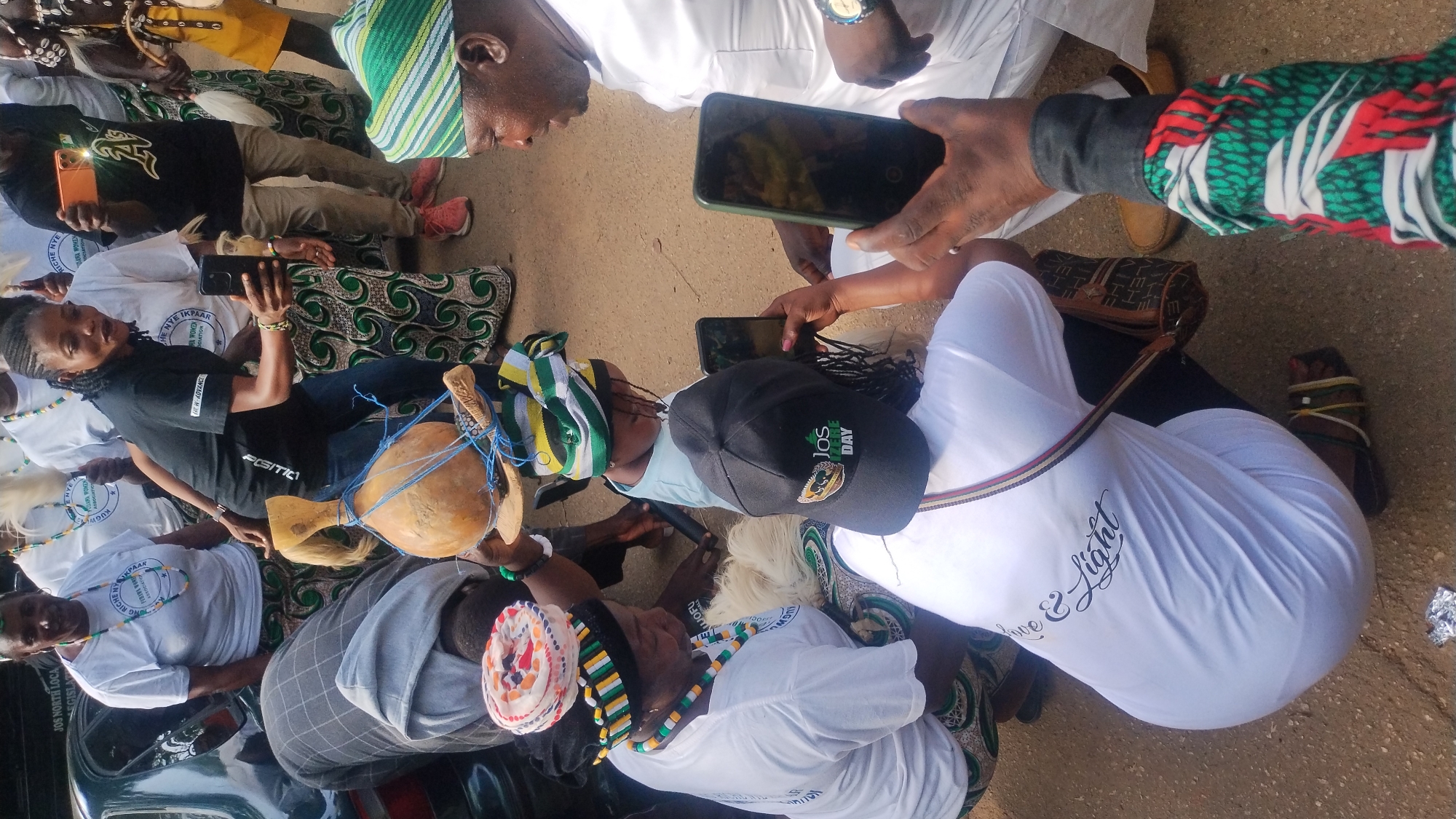 The image depicts a gathering of people outdoors, likely in Jos, Nigeria. Several individuals are wearing traditional attire featuring vibrant green, yellow, and white patterns. Some are also adorned with beaded necklaces and headwear. A gourd instrument is prominently featured, suggesting a cultural or ceremonial event.
The participants appear engaged, with some holding and taking pictures with smartphones, indicating a contemporary aspect to the event. Others are looking at the camera or interacting with each other, suggesting a lively and social atmosphere. The setting appears to be a dusty or paved ground, typical of an outdoor public space. The sunlight suggests it is daytime.
Visible text on clothing and accessories includes "JOS NORTH LOCA", "LEGISLAT", "ASSOCIATION", "RICHE HYE IKPAAR", "DARA WOMEN", "WOFU", "POSITICI", "W-ADVANCE", "DAY", and "Love & Light". These fragments of text provide clues about the nature of the gathering, possibly a cultural festival, a community event, or a commemoration organized by local associations. The presence of both traditional elements and modern technology points to a blend of culture and contemporary life in Jos.