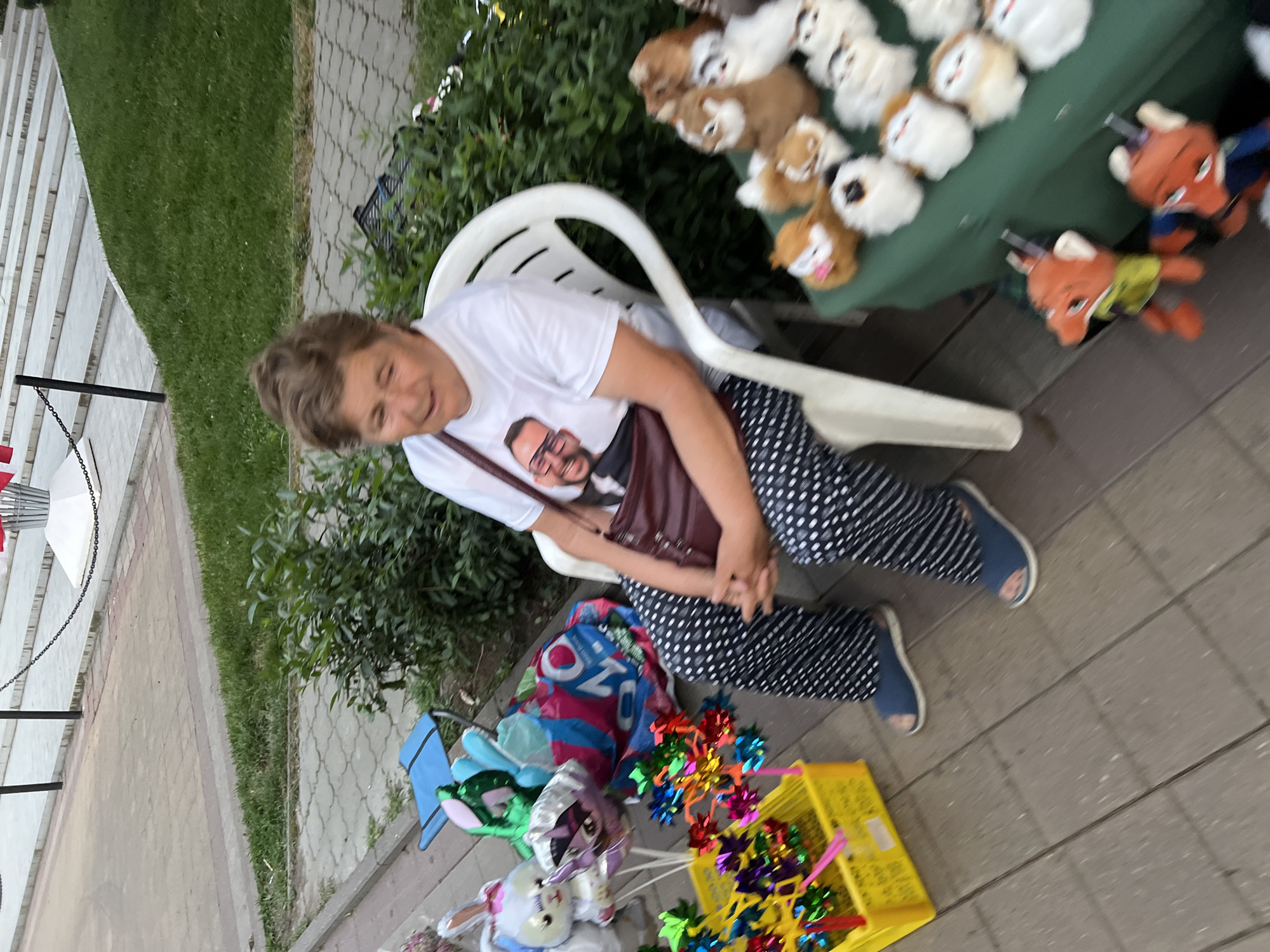 An outdoor scene depicts a woman sitting on a white plastic chair, facing forward with a slight smile and looking down. She is wearing a white t-shirt with a printed image of a man, black and white polka dot pants, and blue open-toed sandals. A dark red purse is slung over her right shoulder. Beside her, to her left, is a yellow plastic basket filled with colorful pinwheels and some balloons, including a white bunny-shaped balloon and a green balloon. To her right, on a green surface, are several small stuffed animals. The background shows a grassy area and a paved walkway with steps, suggesting a public park or pedestrian area. The scene is likely in Krasnodar, Russia, given the context provided.
