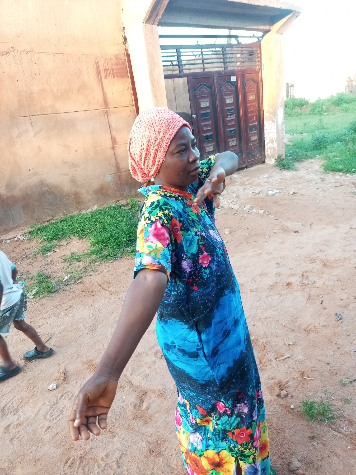 In Tempure, Nigeria, a woman with dark skin is shown from the mid-torso up, looking to her left and extending her right arm. She is wearing a bright pink headscarf and a blue, green, and red floral-patterned dress. Her facial expression is neutral, and her right hand is open and reaching forward, with her fingers slightly curled. In the background, a dirt path is visible, with patches of dry grass and a wall constructed of rough, tan-colored material. To the right of the woman, there's a large, ornate wooden gate, likely part of a building entrance. A hint of greenery and another, smaller gate can be seen beyond the main gate. To the left and slightly behind the woman, the legs of a child wearing shorts and dark clogs are visible, suggesting the presence of another person in the scene. The overall lighting suggests it is daytime, with the sun casting soft shadows.