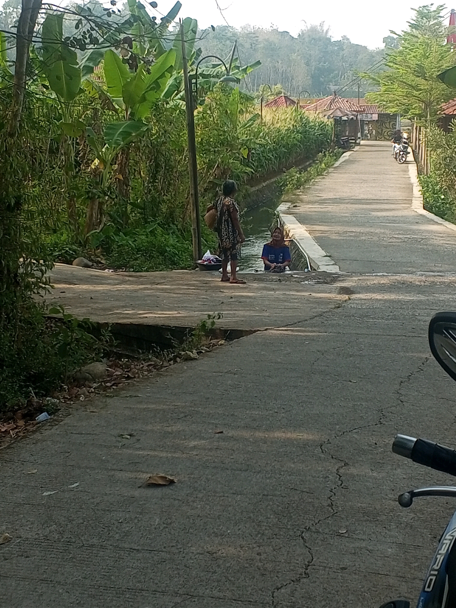 The image depicts a rural scene along a paved road with a drainage canal running alongside it, likely in the city of Sari, Indonesia. Two individuals are present. On the left, a person in a dark patterned dress stands on a concrete bridge spanning the canal, carrying a basket on their back. To the right, in the canal itself, another person wearing a blue shirt is partially submerged, appearing to be washing or playing in the water. Lush green vegetation, including banana trees, lines the canal and the surrounding area. In the background, a motorcycle is visible on the road, and further in the distance, houses with red tiled roofs and lush hills are seen under a hazy sky. A motorcycle handlebar is visible in the foreground on the right side of the image.