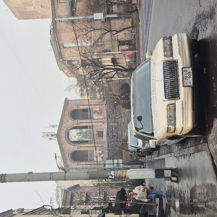 The image depicts an urban street scene in Yerevan, Armenia, specifically featuring the Komitas Chamber Music House in the background. The sky is overcast. In the foreground, a light beige/cream Lincoln Town Car with an Armenian license plate "70 LO 909" is parked on the wet street, angled slightly toward the camera. Behind it, other vehicles are visible. To the left, a pedestrian, likely female, wearing a light jacket and blue jeans, walks away from the camera on a wet sidewalk, carrying a closed black umbrella. A tall utility pole stands on the sidewalk. In the midground, the Komitas Chamber Music House, an orange-pink tuff building with a distinctive three-arched facade on its upper level, is visible. Large banners depicting human figures are affixed to the facade, with a central vertical banner and two smaller horizontal banners. A wide stone staircase leads up to the building. Cellular antennas are mounted on the building's roof. To the right, additional multi-story urban buildings and bare trees are present, along with a red traffic light. The overall environment suggests damp, possibly cold, weather conditions.