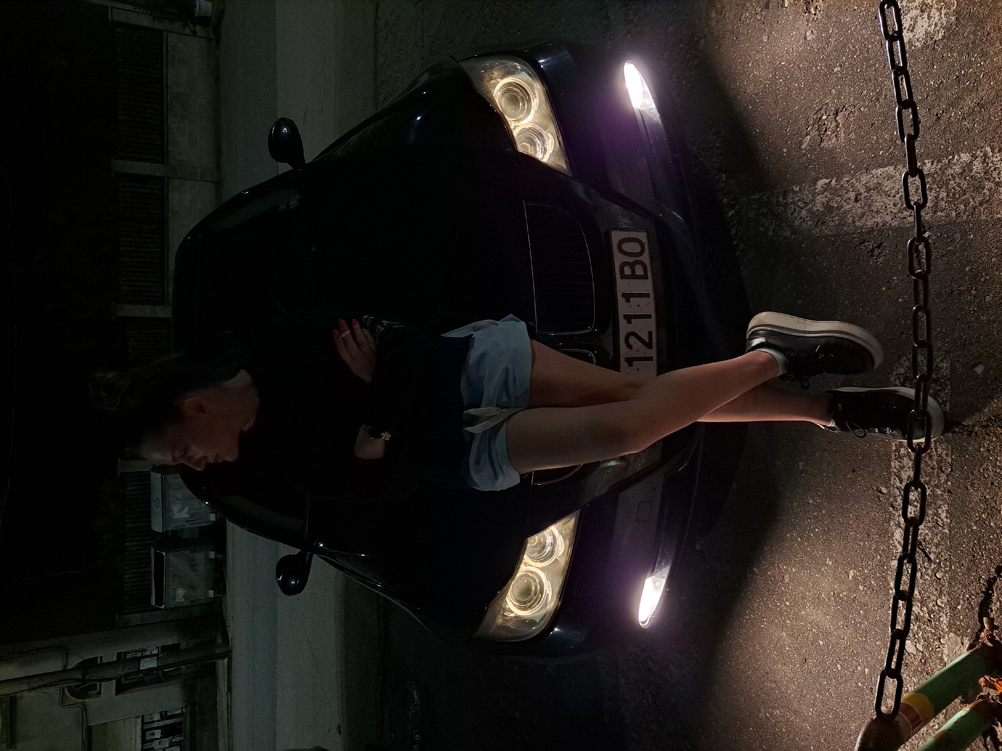 A woman lies across the hood of a black car, with her legs extended towards a curb and chain. The car's headlights are on, illuminating the scene. A license plate is visible on the car's front bumper that reads "1211 BO". The location may be the city of Kaleytsa, Bulgaria. A building is visible in the background, and the surrounding environment appears to be a street or parking area at night. The woman is wearing shorts, a dark top, and platform sneakers. She holds a cigarette.