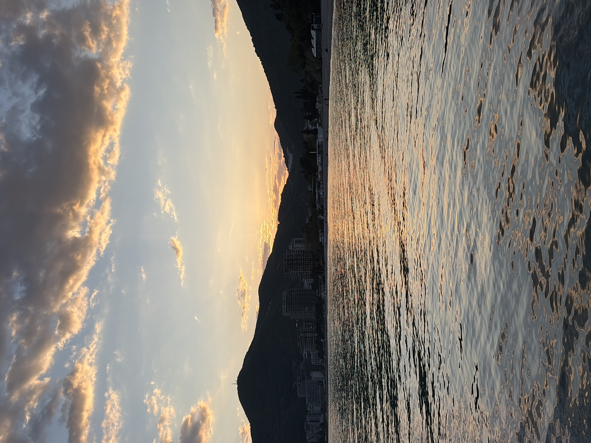 The image depicts a serene sunset over the Adriatic Sea in Budva, Montenegro, featuring a captivating blend of natural landscape and urban development.
In the foreground, the calm surface of the sea stretches across the frame, reflecting the warm, golden hues of the setting sun. The water's surface is textured with gentle ripples, creating shimmering patterns of light and shadow in shades of gold, orange, and deep blue-grey.
The middle ground is dominated by the imposing silhouette of a dark, rugged mountain range that runs horizontally along the coastline. Nestled at the base of these mountains, a coastal settlement or resort area is visible. Numerous buildings, including what appear to be modern high-rises and other structures, are scattered along the shoreline, some featuring exposed frameworks, suggesting ongoing construction or distinct architectural styles. A narrow strip of beach is faintly discernible between the water and the mountain base.
The sky above the mountains is a key feature, showcasing a dramatic sunset. The sun, positioned low on the horizon and partially obscured by the mountains, casts a brilliant golden glow. This light illuminates the underside and edges of scattered clouds, transforming them into soft, golden-orange formations. Further from the sun's direct light, the clouds transition to darker, greyish-blue tones against a lighter blue sky.
The overall scene captures the "golden hour" with its warm, soft light, creating strong contrasts and a tranquil atmosphere. There is no visible text in the image.