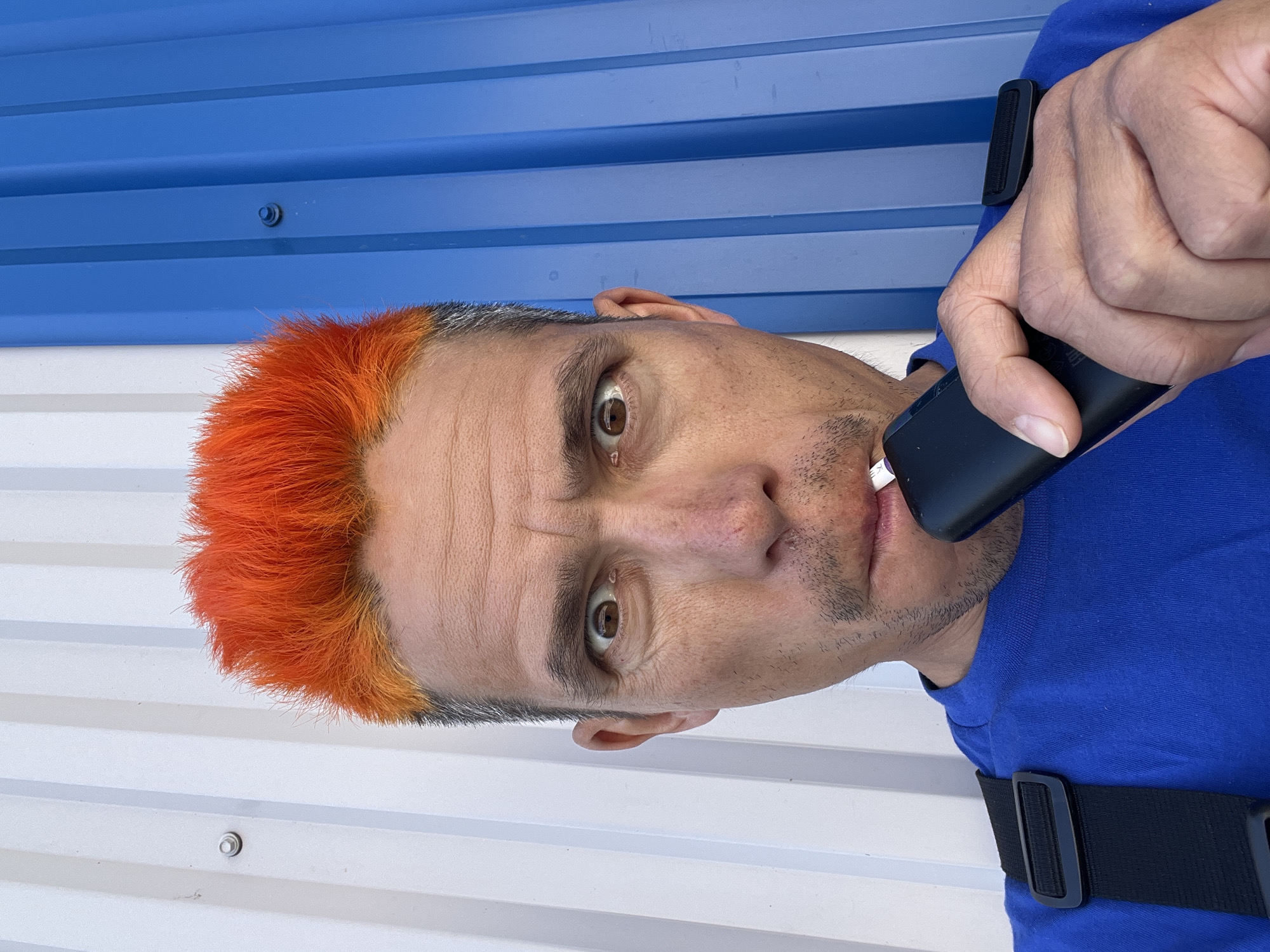 A man with orange hair stands in front of a blue and white corrugated metal wall. He is holding a black device to his mouth. He is wearing a blue shirt with black straps. The location is speculated to be in the city of Chaikovsky, Russia, but there is no definitive evidence in the image to support this claim.