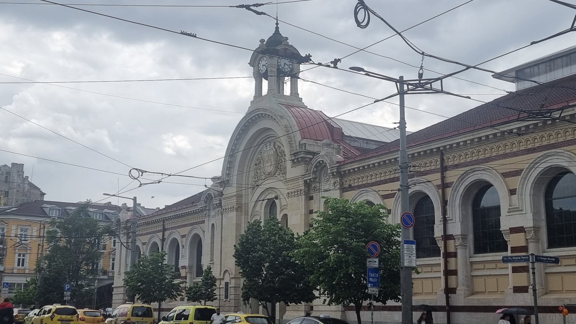 This is a street-level daytime shot of the Central Market Hall in Sofia, Bulgaria. The architectural style is predominantly Neo-Renaissance, featuring a large arched facade with a clock tower atop a red dome. Ornate detailing and arched windows are visible along the building's exterior. Street signs indicating taxi services and street names (e.g., "бул. Кн. Мария Луиза" and "ул. Екзарх Йосиф") are present near a street lamp post. Yellow taxis are parked along the street in front of the building, and pedestrian activity is visible, including individuals with umbrellas. Utility lines are strung across the sky, partially obscuring the cloudy sky above. Other buildings are visible in the background across the street.