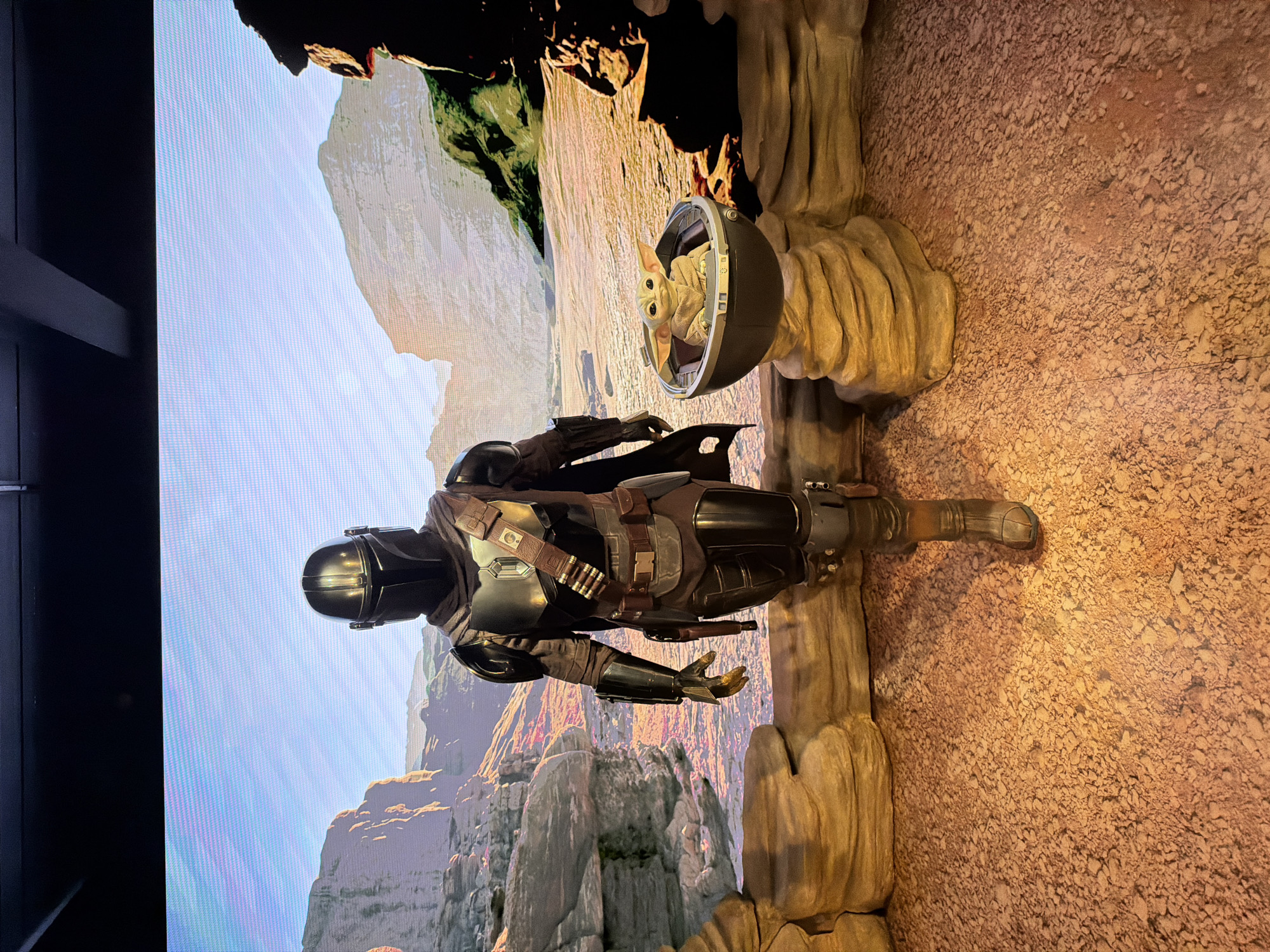 A life-sized statue of the Mandalorian and Grogu is displayed in front of a large screen showing a rocky landscape. The Mandalorian stands on a beige, rocky display, with his right arm extended. Grogu is in his carrier. The scene simulates the environment of the fictional planet of Arvala-7 from the Star Wars TV series, "The Mandalorian." The location is unknown, as no specific markers indicate London, UK.