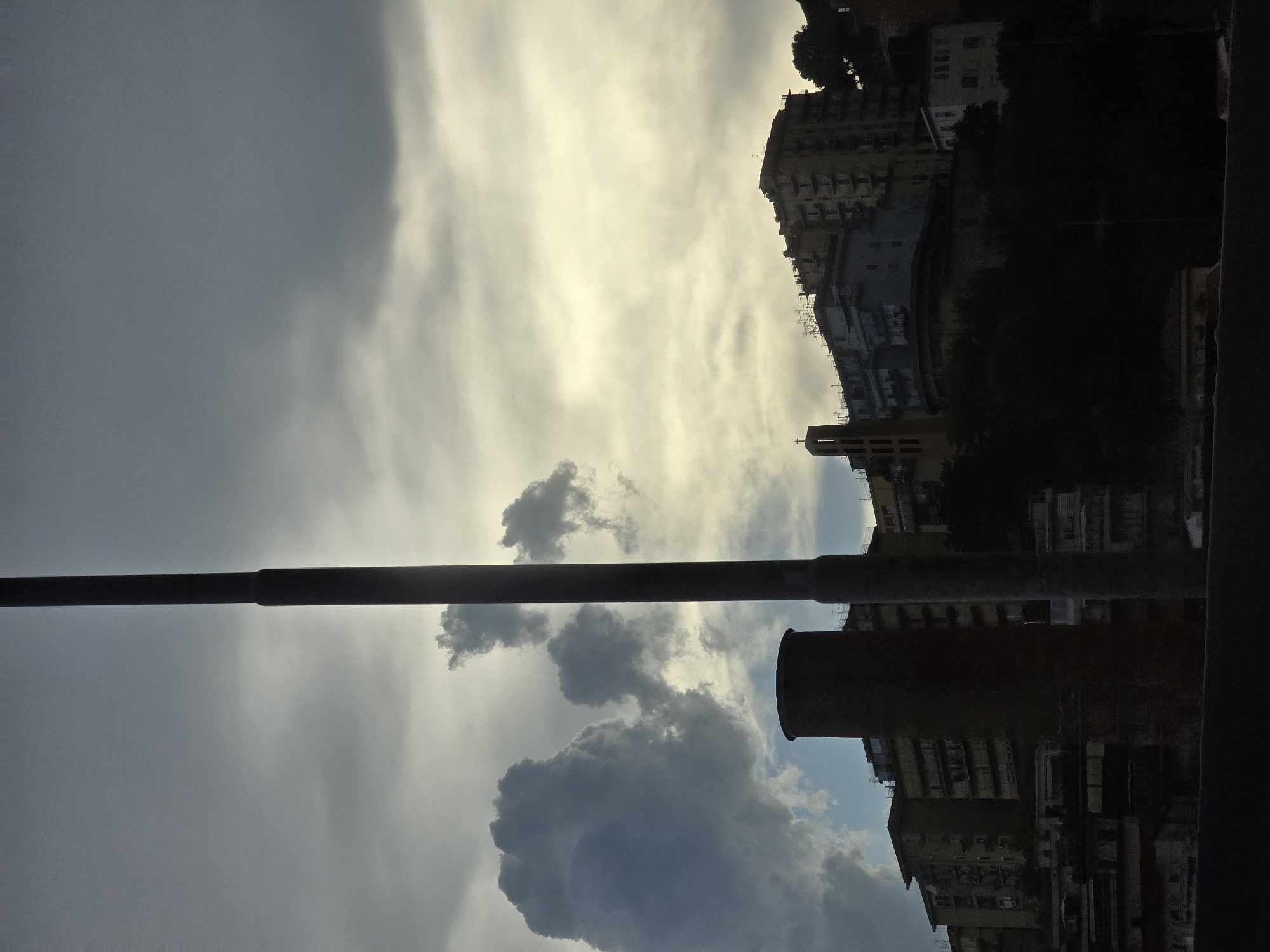 The image captures a dramatic sky over the city of Salerno, Italy, as evening approaches. A tall, dark lamppost dominates the foreground, its silhouette stark against the turbulent clouds. To the left, a large, dark cumulus cloud formation looms, contrasting with patches of lighter, yellowish sky peeking through elsewhere. In the mid-ground, a circular brick structure, possibly part of an industrial building or a historical tower, stands prominently. Behind it, a series of multi-story buildings climb the hillside, their facades illuminated by the dimming light. A slender, steeple-like structure, likely a church tower with a cross at its apex, is visible among the buildings. The lower portion of the image is heavily shadowed, obscuring many details of the urban landscape and suggesting that the viewpoint is from a lower elevation, looking up towards the city perched on the hill. No people are visible in the frame.