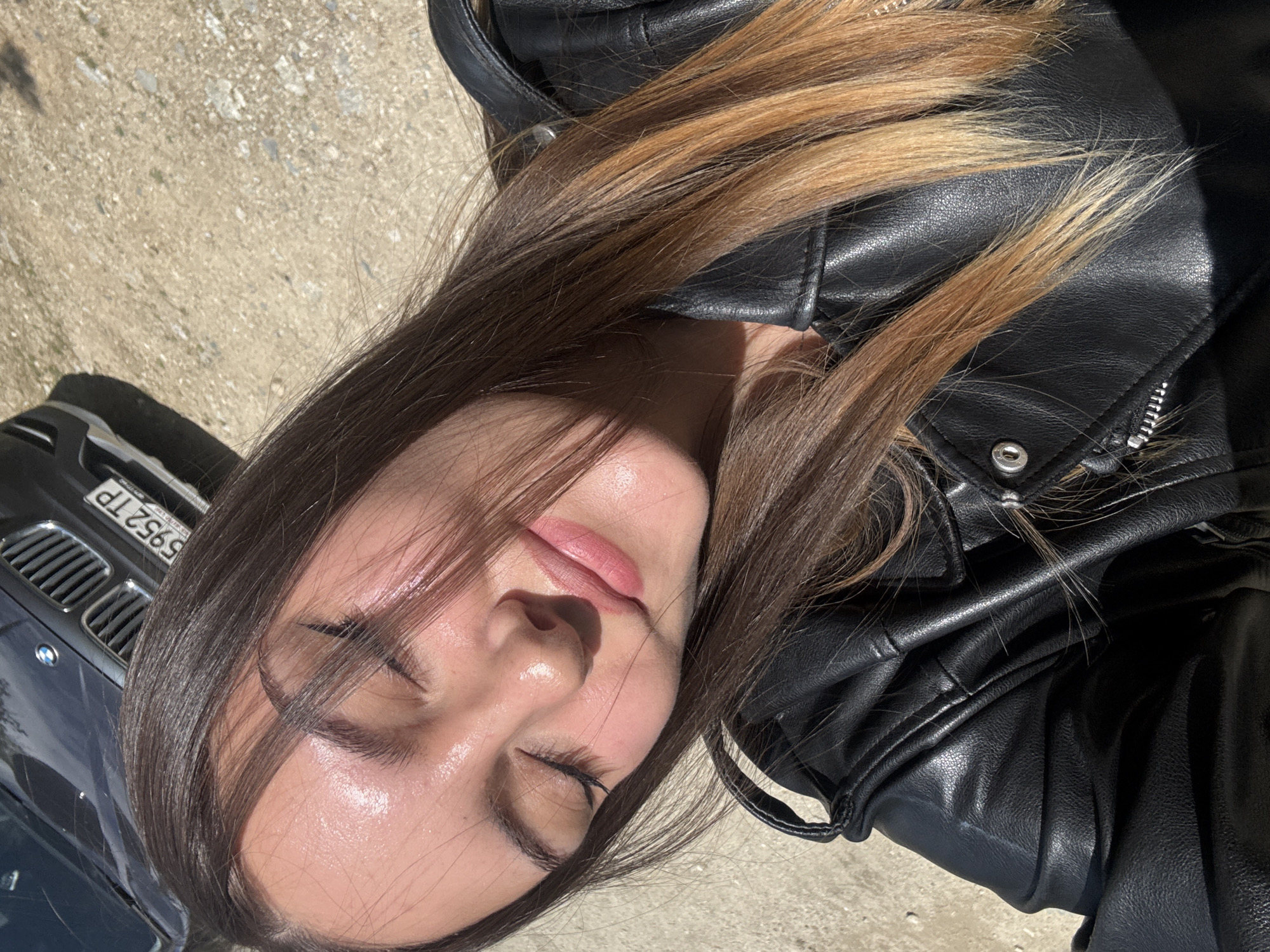 A young woman lies on a concrete surface with a black leather jacket draped around her upper body. She has long, dark brown hair with light brown highlights. Her eyes are closed, and her face is turned towards the sun. A dark-colored BMW car is visible to the left of the frame, with the license plate "5952TP." The setting appears to be outdoors, likely in a public area within the city of Ovchartsi, Bulgaria. The lighting suggests it is daytime with bright sunlight.