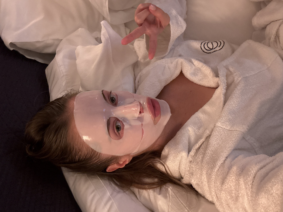 A woman lies on a bed covered with white bedding, wearing a white bathrobe. She has a sheet mask applied to her face. Her head is tilted to the side, and she's looking directly at the camera. Her right hand is raised in the air, fingers spread. The bathrobe has a simple logo on the chest. The scene is lit with a soft, warm light, creating a cozy atmosphere.