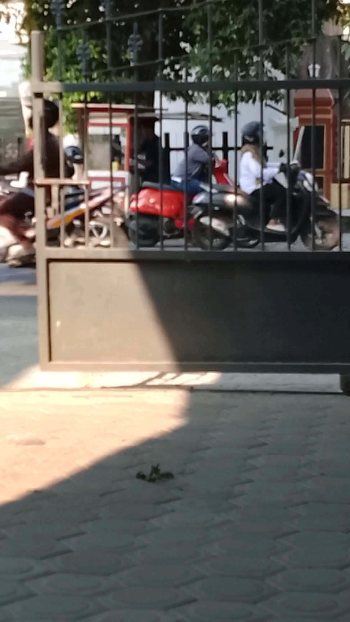 The image depicts a street scene in Slawi, Indonesia, characterized by a metal gate in the foreground casting a diagonal shadow across a patterned tiled surface. Beyond the gate, several motorcycles are visible, some with riders. In the mid-ground, two individuals wearing helmets are seated on motorcycles. The person on the left is on a red scooter, while the person on the right is on a black scooter. They are both facing away from the camera, and appear to be waiting or in motion. The background shows greenery and what appears to be a building structure. The overall lighting suggests it is daytime, with strong sunlight creating distinct shadows.