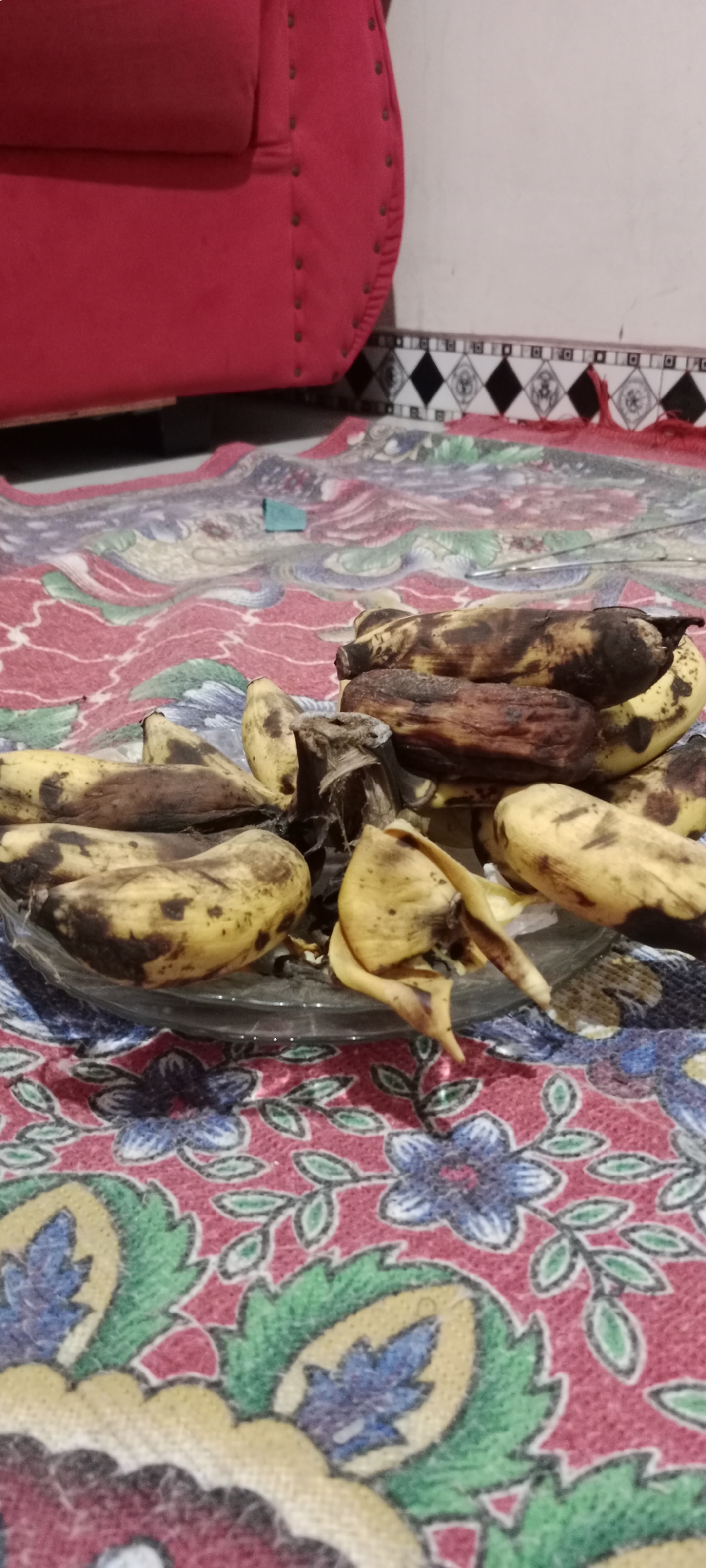 A pile of overripe bananas sits on a clear glass plate on top of a red, floral-patterned cloth. The bananas are yellow with significant brown and black spots, indicating a high level of ripeness. Some of the bananas appear to be partially peeled. In the background, a portion of a red sofa is visible, as well as a wall with a decorative border. The overall setting suggests an indoor environment, possibly a home. No people are present in the image. The location is specified as Soko, Indonesia.