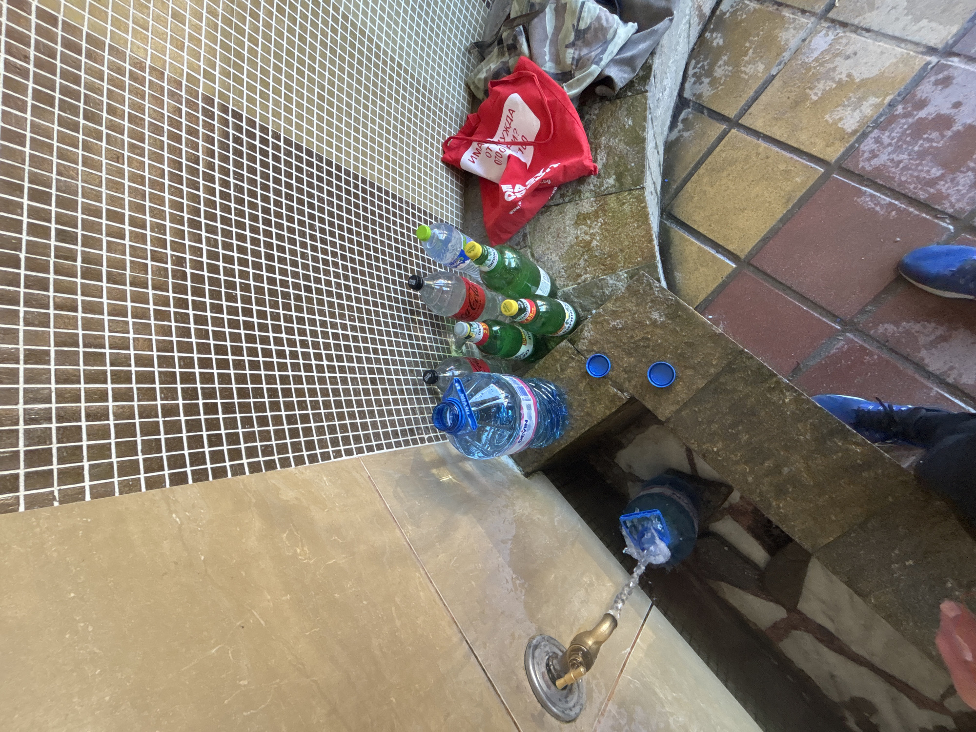 The image presents an outdoor scene, likely a public area in the city of Zlatograd, Bulgaria, as indicated by the inscription on a red bag. The scene is dominated by a water source; a metallic spigot with water flowing out. Around the spigot, there is a collection of various plastic bottles, some empty and some full. There are plastic bottles of water and soda, and a red reusable shopping bag. The ground is tiled in different patterns; a brown tiled wall is also visible. A pair of blue shoes and part of a black garment are visible in the upper-right.