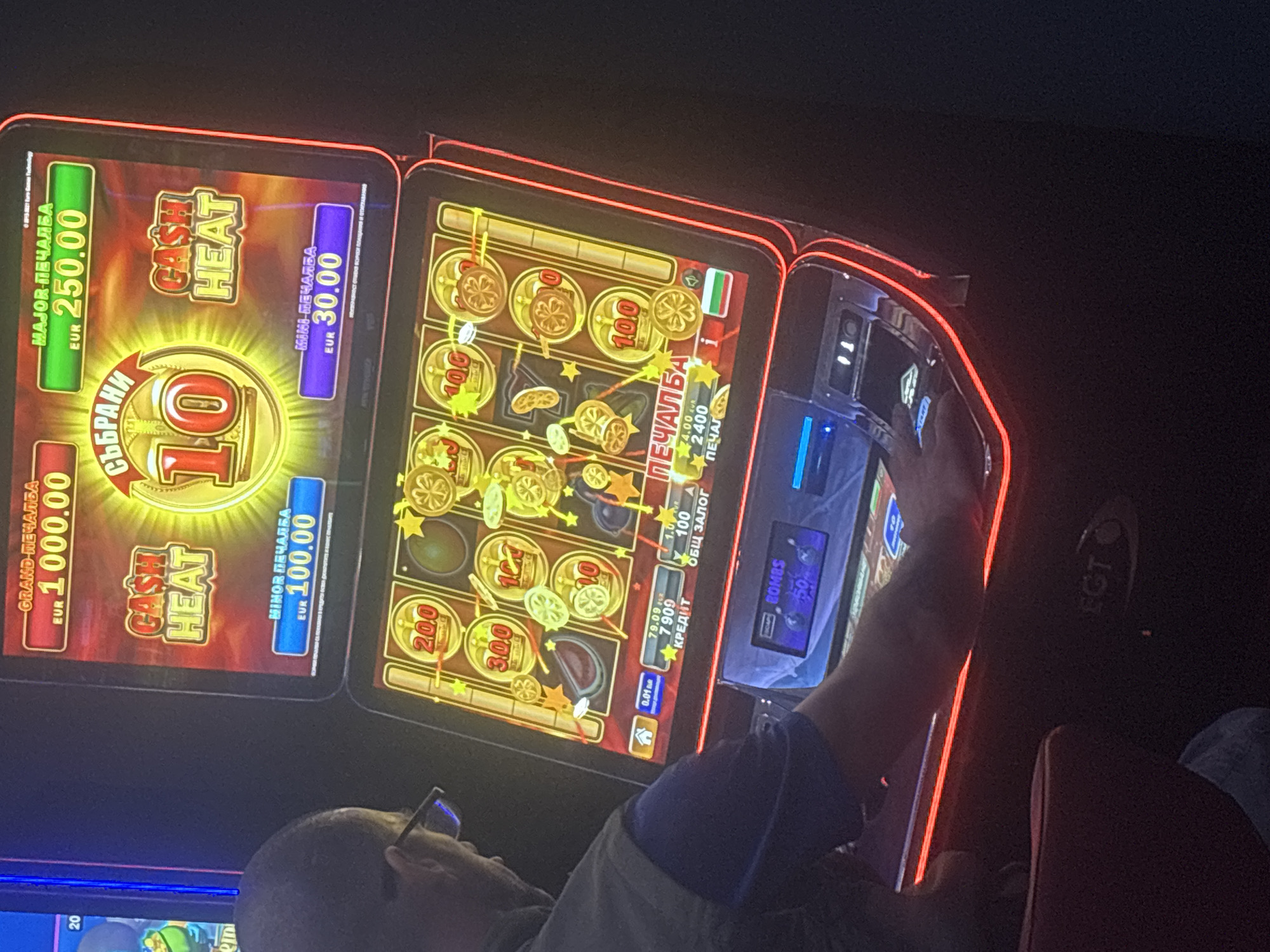A man is playing a slot machine in a dimly lit casino in Beloslav, Bulgaria. The slot machine has a red and gold color scheme with various flashing lights and game displays. The man is wearing glasses and a blue shirt with a grey vest. He is seated and appears to be concentrating on the game. The slot machine features text in Bulgarian, including "GRAND PEЧАЛБА" (Grand Win) and "MAJOR PEЧАЛБА" (Major Win). There are also displays showing monetary values like "1000.00 EUR" and "250.00 EUR". The bottom part of the machine shows game credits, total bet, and other game-related information, including "BOMBS" with a value of "50". The EGT logo is visible on the lower part of the machine.