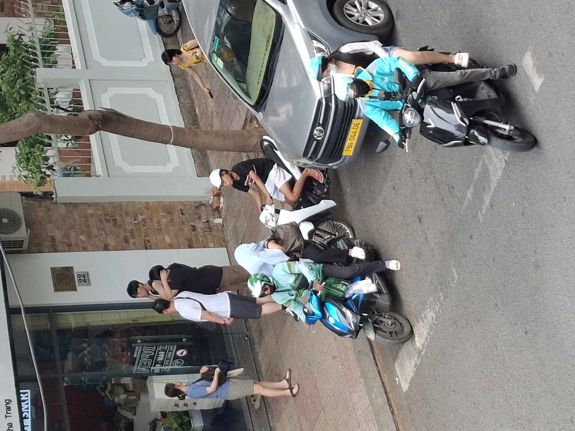The image depicts a street scene in Nha Trang, Vietnam, likely near a commercial establishment. In the foreground, three motorbikes are visible. The leftmost motorbike is a blue and black scooter, with a person wearing a green jacket and helmet riding it, and another person wearing a light blue head covering and dark clothing as a passenger. The middle motorbike is a white scooter, with a person in a black t-shirt and white shorts, wearing a white helmet, riding it. The rightmost motorbike is a black scooter, with a person in a teal jacket and dark pants, wearing a teal helmet and black mask, riding it, and another person in a white shirt and denim shorts, wearing a teal helmet, as a passenger. A silver car is parked between the middle and rightmost motorbikes. On the sidewalk to the left, several people are standing. Two individuals, one in a blue shirt and shorts, the other in a white t-shirt and shorts, are near the entrance of a shop with a glass facade. Two other individuals, one in a black t-shirt and the other in a black shirt, are standing near them. The shop appears to be a retail store, with displays visible through the window. A tree with a white band around its trunk grows on the right side of the image, near a white fence. The environment suggests an urban setting with paved roads and sidewalks.