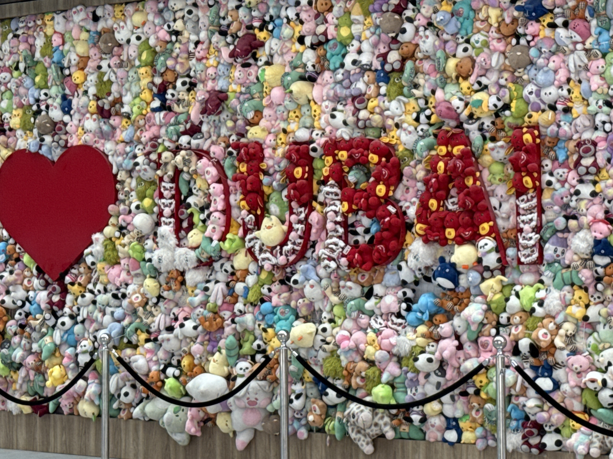 The image displays a vibrant wall covered entirely with an assortment of stuffed animals. The stuffed animals are predominantly in pastel and bright colors, including pink, yellow, green, blue, and white, creating a playful and chaotic aesthetic. Prominently featured in the center of the wall are large, red, three-dimensional letters spelling out "DUBAI". To the left of the letters is a large, bright red heart shape. Two stanchions with black ropes are positioned in front of the wall, acting as a barrier. The stuffed animals are densely packed, with some appearing to be stacked upon each other, and they vary in size and type, including bears, rabbits, and other animal characters. The scene appears to be an outdoor installation or a wall within a public space in Dubai, United Arab Emirates. There are no people visible in the image.