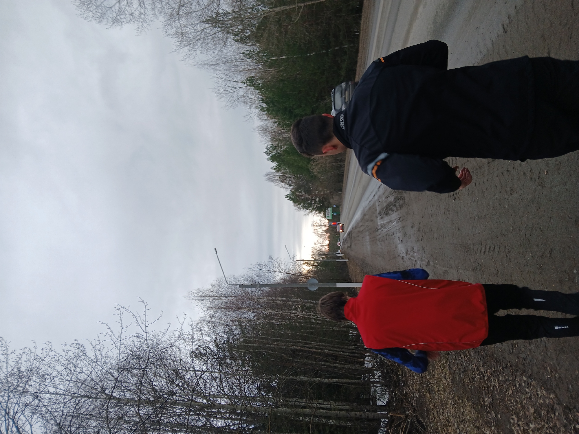 Two individuals are walking alongside a road near what appears to be a wooded area. The person closer to the camera is wearing a red and blue jacket and black pants. The other person is wearing a dark jacket with orange accents. They are walking along a dirt shoulder. The road is a two-lane asphalt road with a car visible in the distance. The sky is overcast. Bare trees are visible along the roadside. The location is in a rural area near the city of Chaikovsky, Russia.