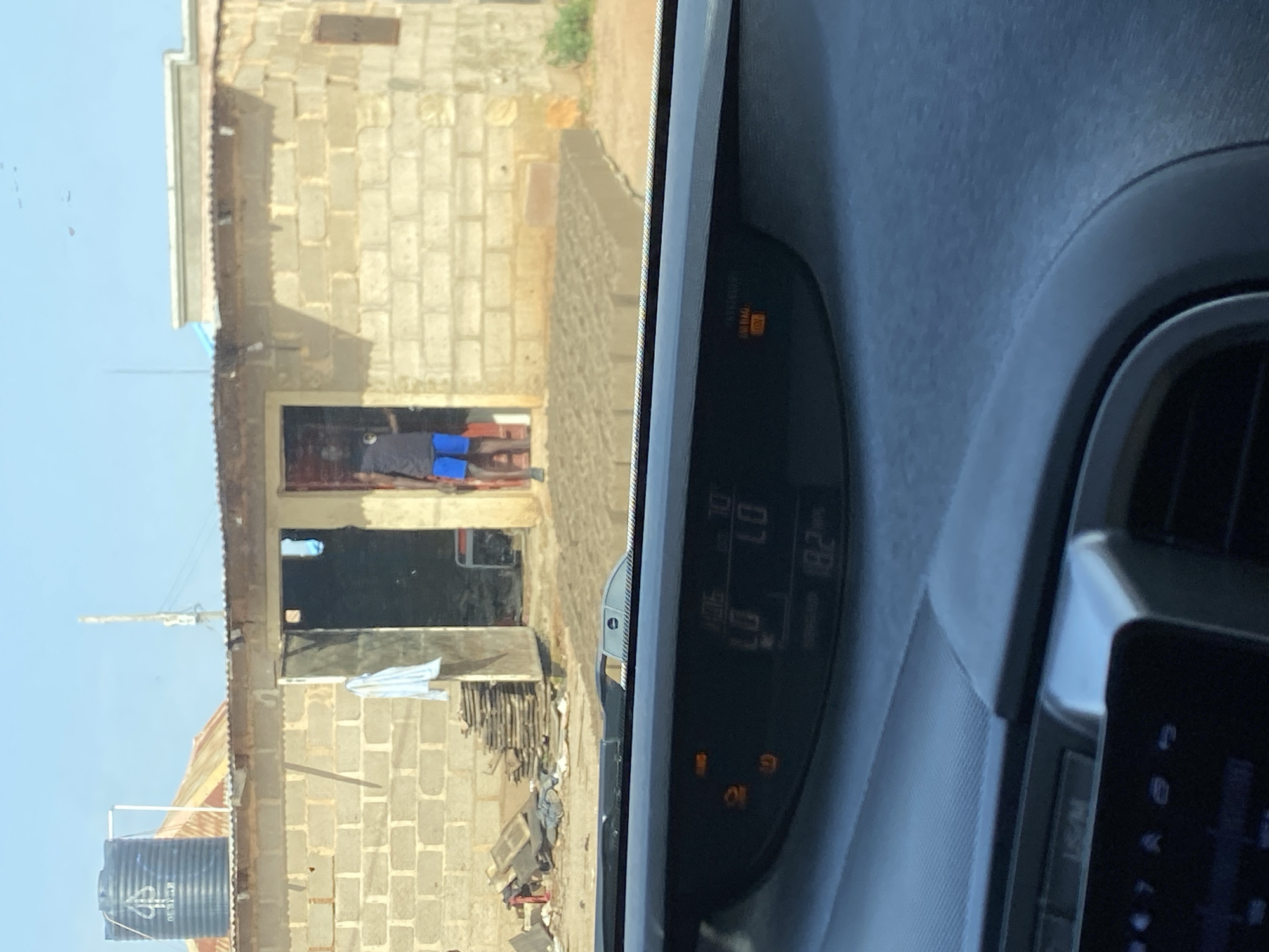 The image captures a street view in Bar Jirgi Summa, Nigeria. In the foreground, the dashboard of a vehicle is visible, displaying various indicators and numerical readouts. The exterior scene shows a building constructed from concrete blocks, with a corrugated metal roof. A person wearing a dark t-shirt and blue shorts stands in the doorway of the building. Adjacent to the building, there are stacks of wooden planks and building materials. A large black water tank is mounted on the roof of a structure to the left. The environment appears to be outdoors, under a clear sky.