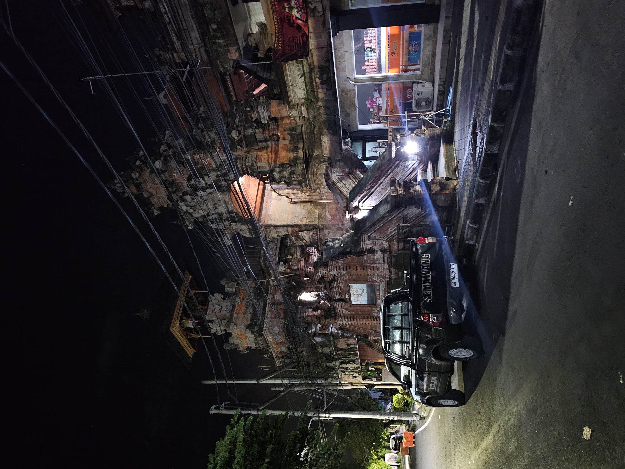 The image depicts a night scene in Denpasar, Indonesia. A black pickup truck with the word "SEMAWANG" on the side and a license plate partially visible as "DK 8606 EG" is parked on the right side of the frame. The truck is positioned near a sidewalk. To the left of the truck is a Hindu temple or traditional Balinese structure adorned with intricate carvings, lit with warm, reddish-orange light emanating from within its arched entrances. Above the temple and on the left side of the frame is a dense network of electrical wires. Next to the temple, further down the street, there is a building with lit-up windows, suggesting a convenience store or small shop, with visible advertisements or signage. Street lighting illuminates the road and surrounding areas. The foreground on the right side shows an asphalt road surface. Trees and other greenery are seen to the left. Overall, the image captures a blend of traditional Balinese architecture and modern infrastructure in an urban night setting.