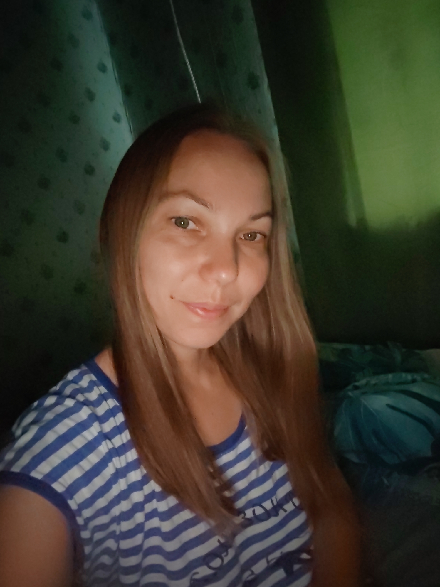 A young Caucasian woman with shoulder-length blonde hair is posing for a selfie in a dimly lit room. She is wearing a blue and white striped t-shirt. Her face is partially illuminated by the camera's flash, revealing her fair skin, green eyes, and a slight smile. A mole is visible on her left cheek. The background is dark and appears to be a room, with a green-tinted wall or curtain to the right and a patterned dark fabric to the left. The overall impression is of an indoor self-portrait taken in low light conditions.