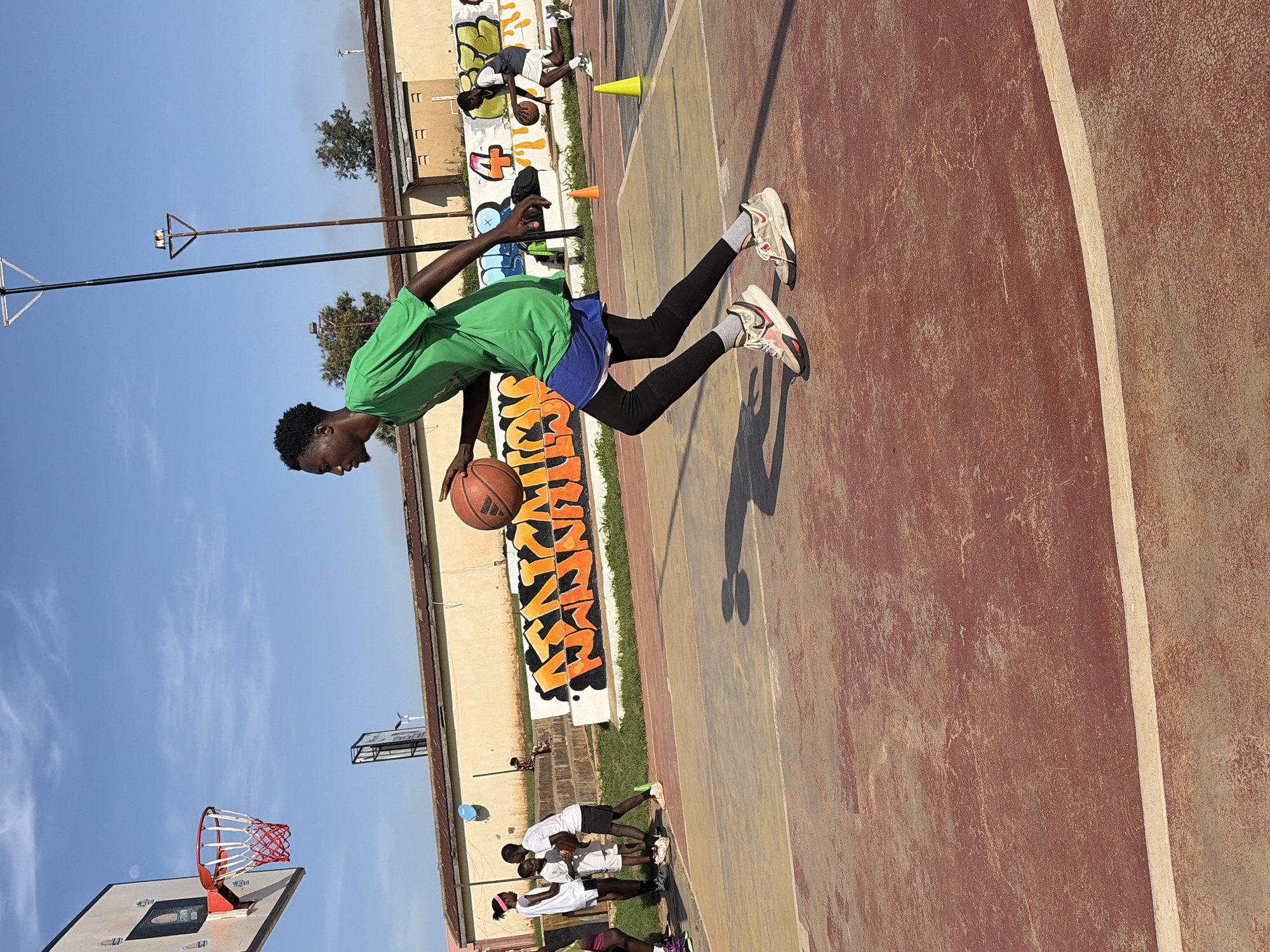 In Jos, Nigeria, on a sunlit day, a group of young people engages in a basketball practice session on an outdoor court. The court's surface is a reddish-brown color, marked with off-white lines. In the foreground, a male teenager, wearing a vibrant green t-shirt, blue shorts, black leggings, and athletic shoes, dribbles a basketball with focus. His body is angled in motion, suggesting he is in the midst of a drill or game.
In the middle ground, several other individuals are visible. To the left, a female and two males, dressed in white attire, appear to be observing or participating. Further back, another male teenager, in a grey shirt and white shorts, is also dribbling a basketball, indicating the activity is widespread.
The background features a light-colored wall adorned with colorful graffiti. Beyond the wall, trees and buildings are visible, under a clear blue sky with some light cloud cover. Two tall poles, likely for lighting, stand on the court, and orange and yellow traffic cones are placed as markers. The overall atmosphere is one of energetic recreation within a community space.