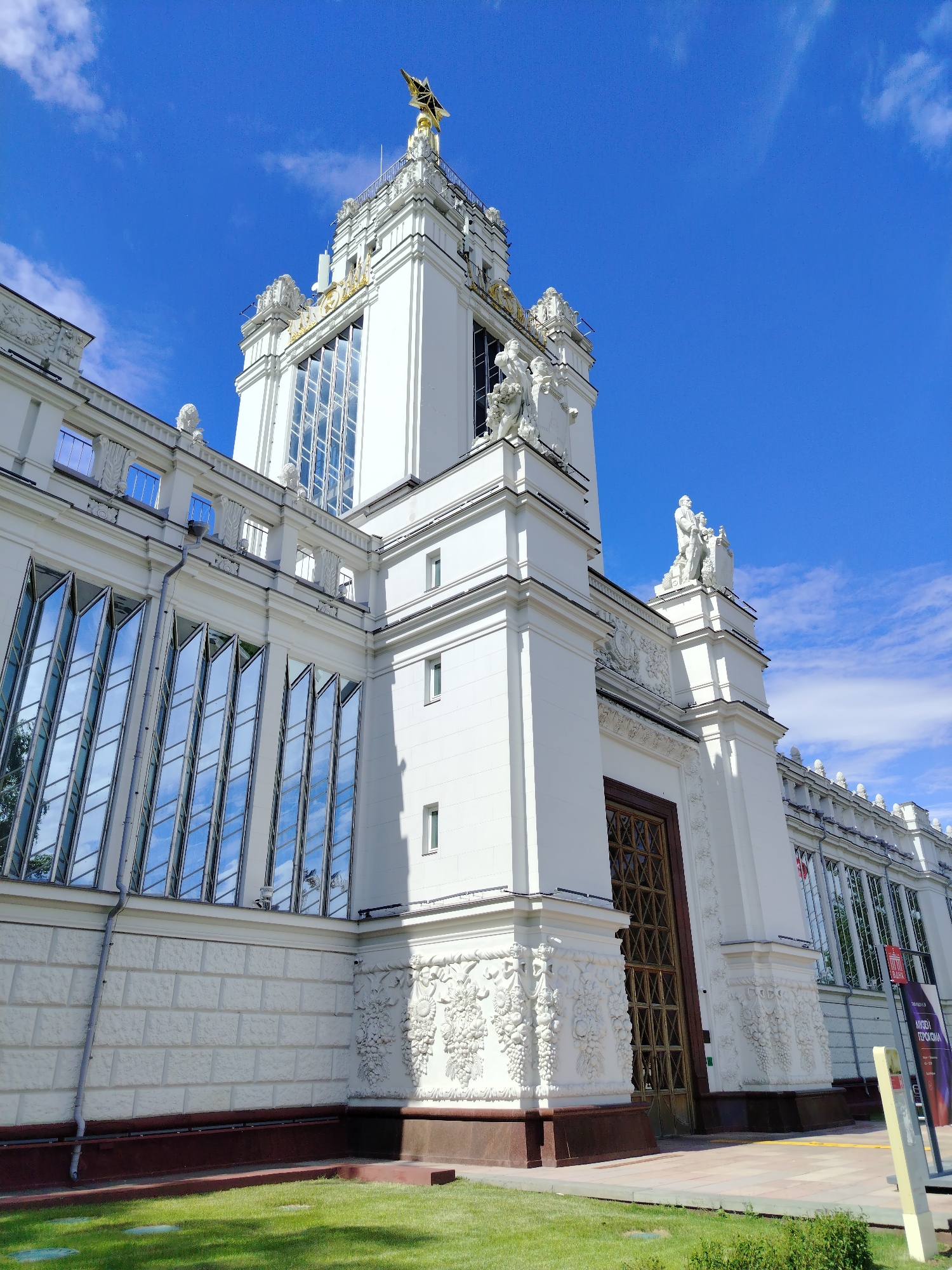 This media file captures a view of a large, ornate white building under a clear blue sky with scattered white clouds. The building exhibits classical architectural elements, including decorative friezes, sculptures, and large, multi-pane windows. A prominent feature is a tall tower topped with a golden star-like emblem. Statues of human figures are visible on ledges and atop the building. The windows on the lower section of the building are angled and reflect the sky. The ground in front of the building is covered with green grass. The overall scene suggests a sunny day, likely in the daytime, given the bright illumination and clear sky. The architecture and the presence of statues suggest this might be a public building or monument in Moscow, Russia, possibly a museum, exhibition hall, or administrative center, given the style often found in Soviet-era architecture. There is some text visible on a sign in the lower right corner of the image, but it is too blurry to read.