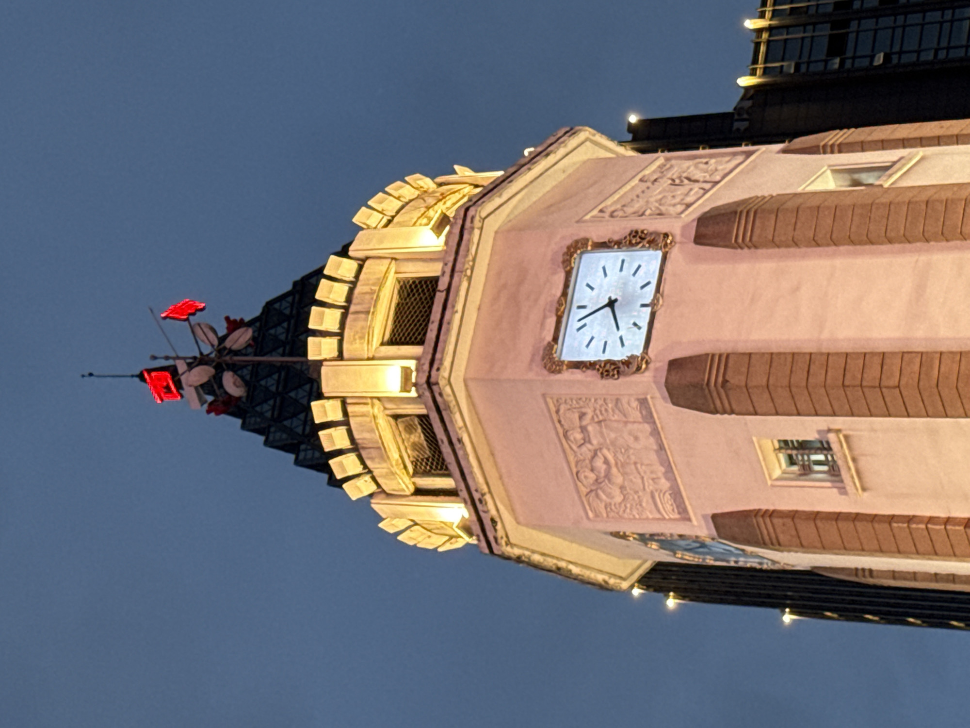 The image shows the Jiefang Monument clock tower in Yuzhong District, Chongqing, China. The tower is illuminated by warm lights, highlighting its ornate facade and the clock face. The clock shows the time is approximately 4:48. Red flags and what appear to be weather vanes are visible at the very top of the tower, against a twilight sky. The tower is surrounded by other buildings, some of which are also lit, suggesting an urban nighttime setting. The architecture of the clock tower appears to be a blend of classical and art deco styles.