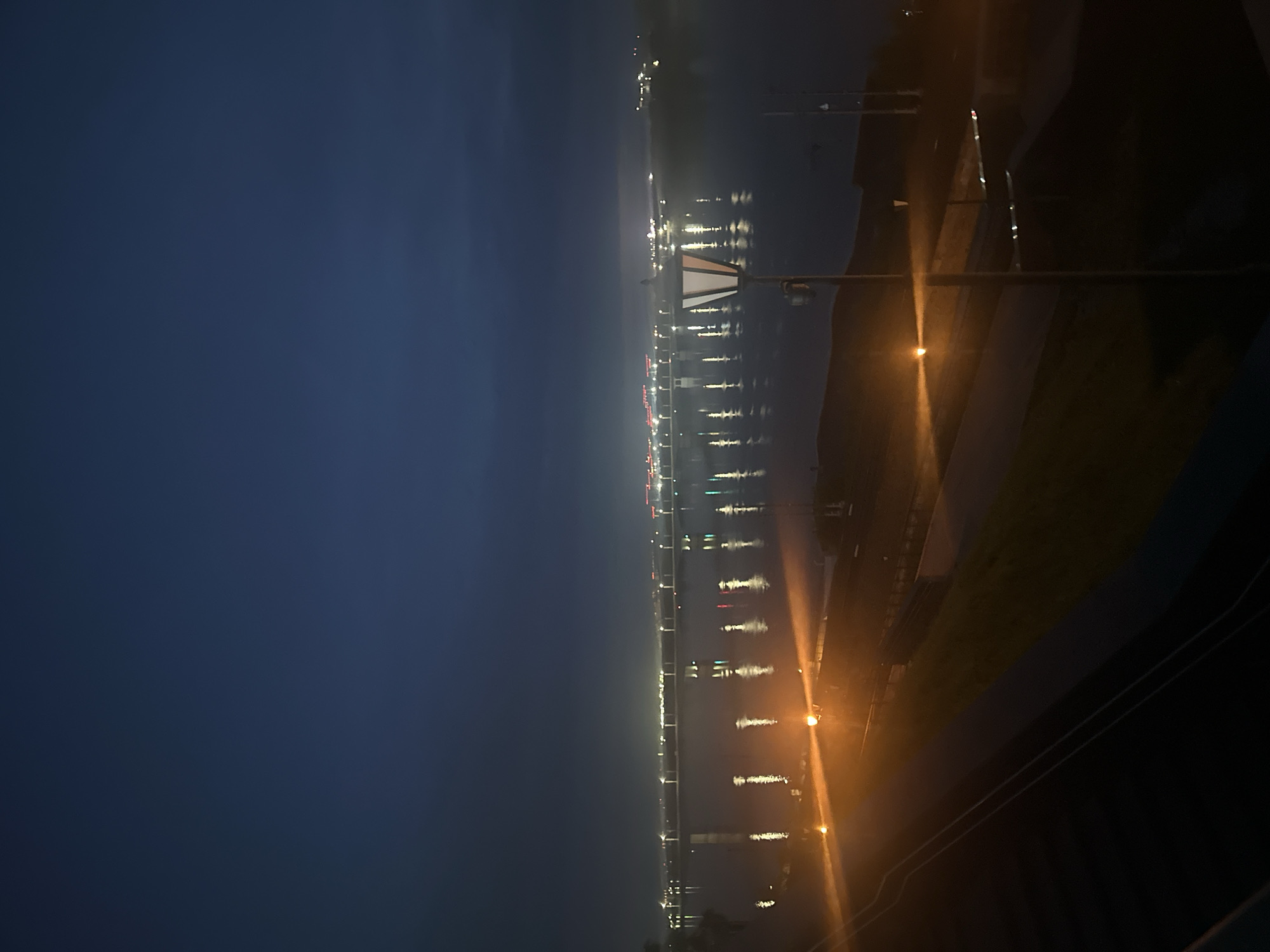 The image shows a nighttime view of the Kama River in Perm, Russia. The skyline on the opposite bank is visible with city lights reflecting in the water. A street light in the foreground provides additional illumination. The sky is dark and overcast. The perspective suggests the photo was taken from an elevated position, possibly a building or high vantage point. The angle and composition provide a panoramic view of the river and city.