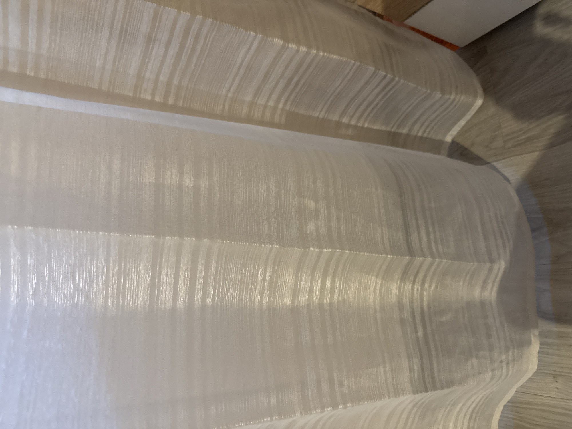 A close-up shot shows a white, translucent, patterned fabric draped in folds. The fabric appears to be smooth and lightweight, with a subtle vertical texture. Behind and above the white fabric, a beige-colored surface is visible, also with a textured pattern. To the right of the image, what seems to be a wood-patterned surface can be seen. The composition is tightly framed, focusing on the textures and patterns of the materials. The location, Lovech, Bulgaria is not determinable from the image.
