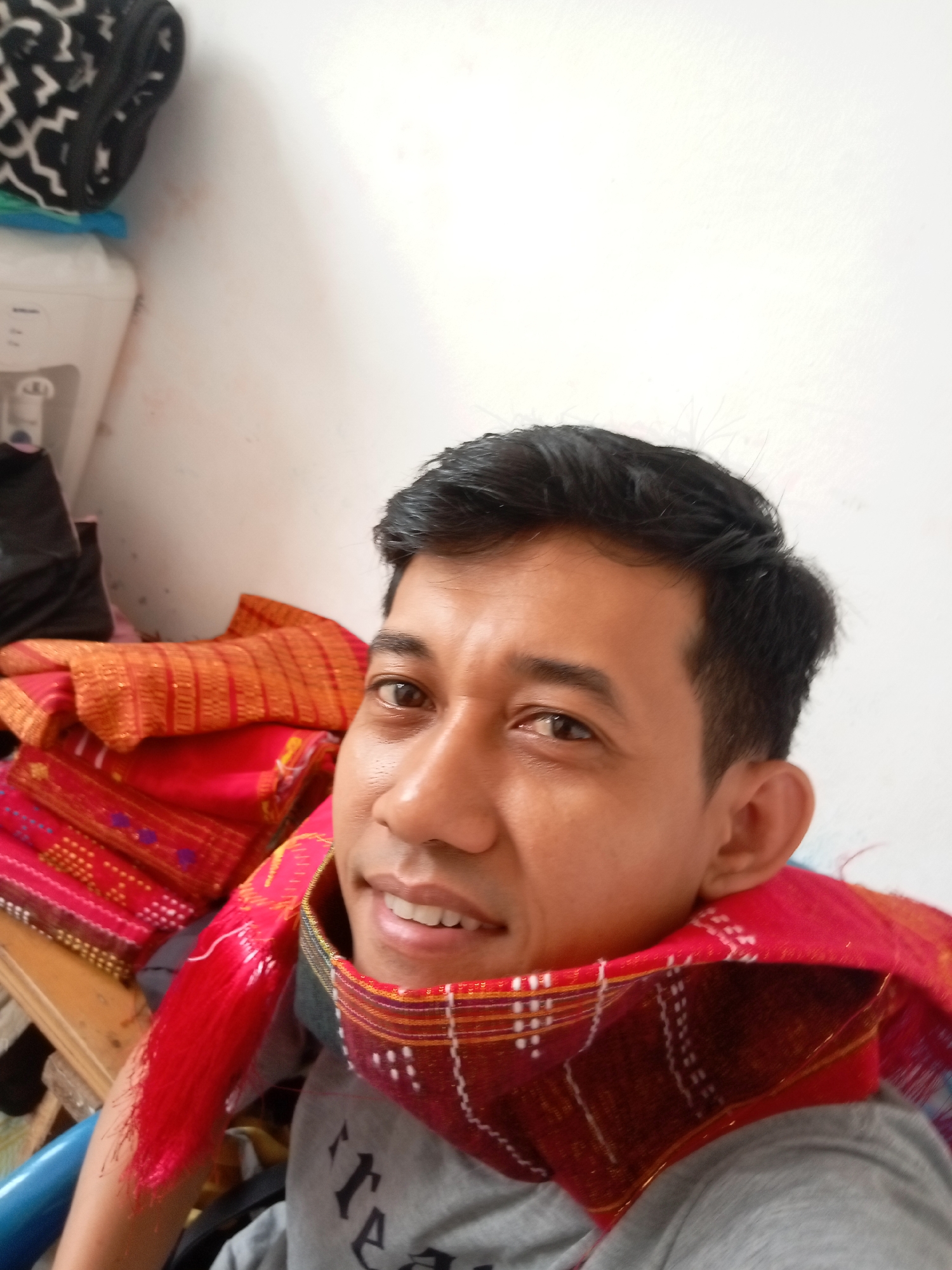 A man is shown from the chest up, with his head tilted slightly to his left and looking at the camera. He has short, dark hair and brown eyes. He is wearing a grey t-shirt and a red and orange patterned scarf around his neck. Behind him, to his left, are stacks of fabric, primarily red and orange with intricate patterns. To the far left of the image, a white appliance with a grey top is partially visible, and above that, a black and white patterned object is perched on top. The background is a plain white wall. The image was taken in Batam, Indonesia.