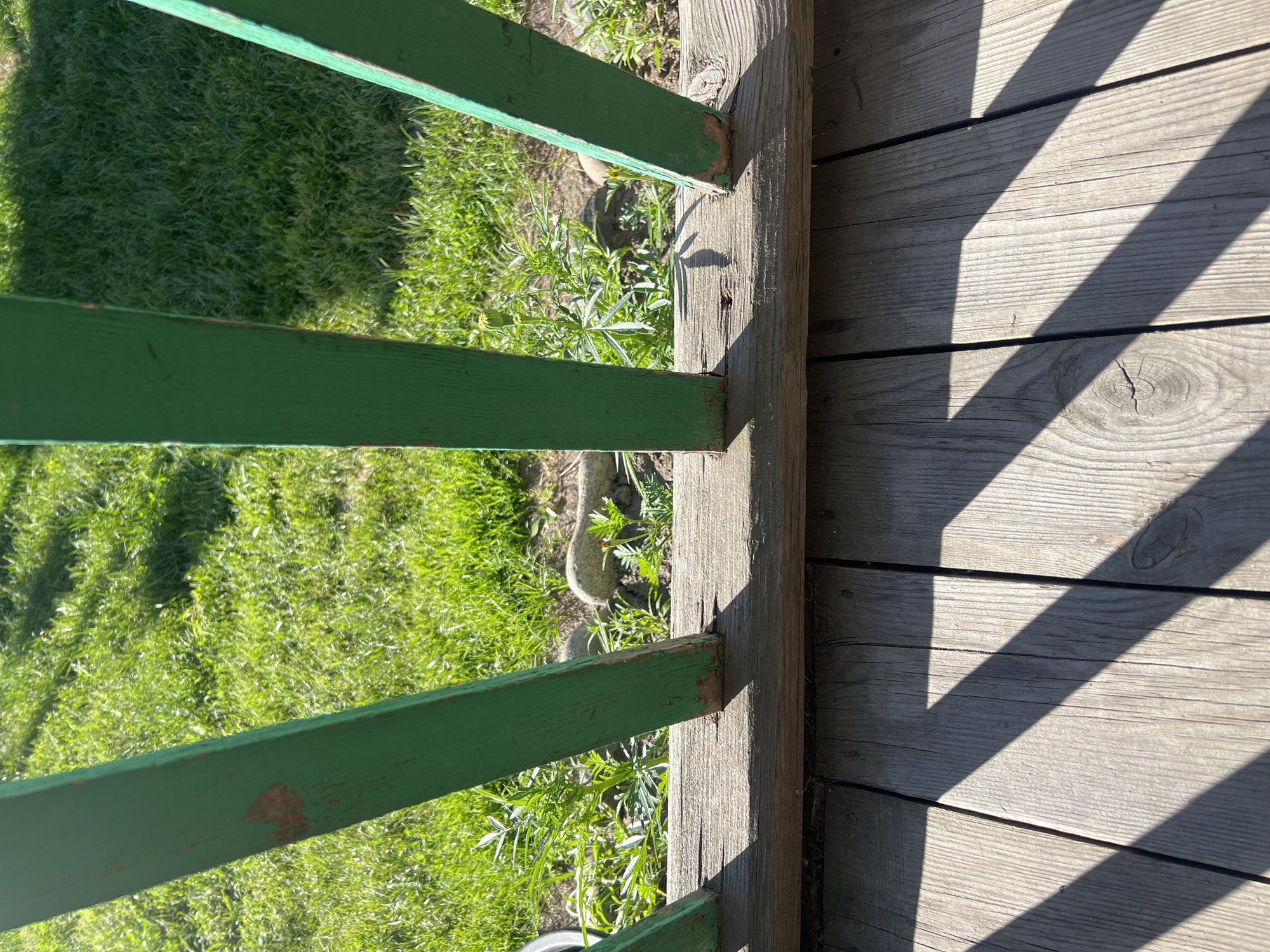 This image shows a close-up view of a wooden porch railing and deck. The railing consists of several vertical green wooden posts, some of which show signs of wear and peeling paint. These posts are attached to a horizontal wooden beam that runs along the top of the deck. The deck itself is made of weathered wooden planks. In the background, green grass and some low-lying plants are visible, suggesting an outdoor setting. The lighting indicates it is daytime, with sunlight casting distinct shadows from the railing onto the deck. There are no people or animals visible in the image. The location is not identifiable as Mochische, Russia, or any specific city.