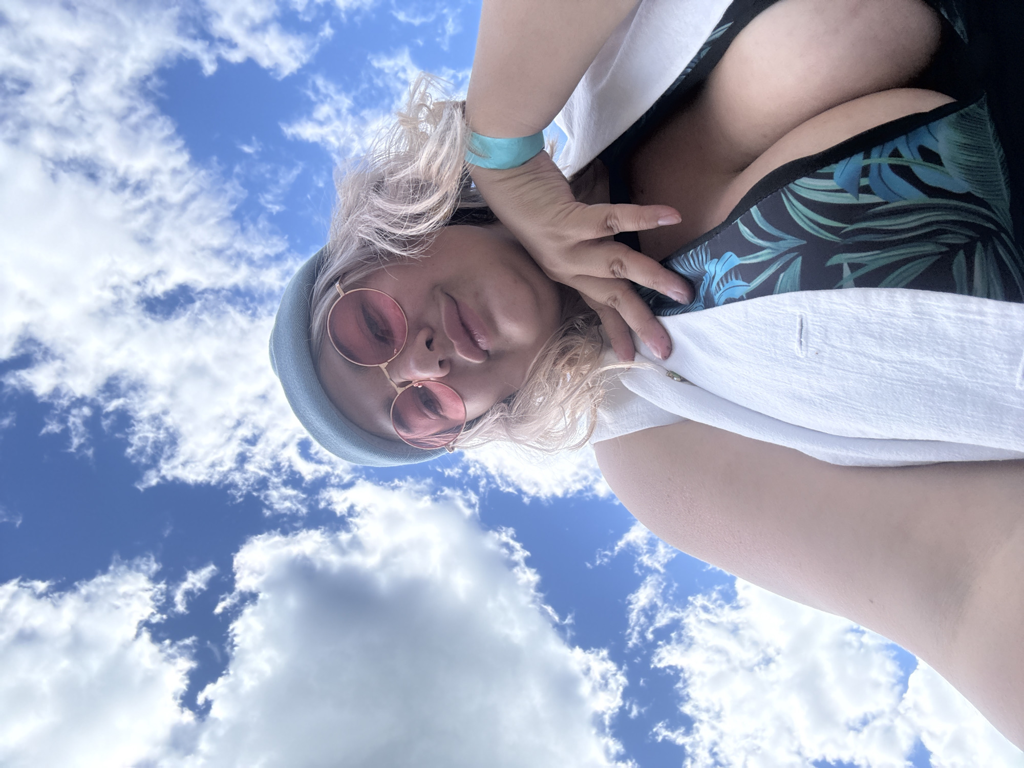 The image is a selfie taken outdoors against a blue sky with scattered clouds. A person is visible from the chest up, wearing a light blue beanie, pink-tinted sunglasses, and a white open vest over a dark swimsuit with a tropical leaf pattern. They are resting their chin on their hand, which is adorned with a teal wristband. The perspective is looking upwards at the person, with the sky occupying most of the background. There is no indication of the city of Birobidzhan, Russia.