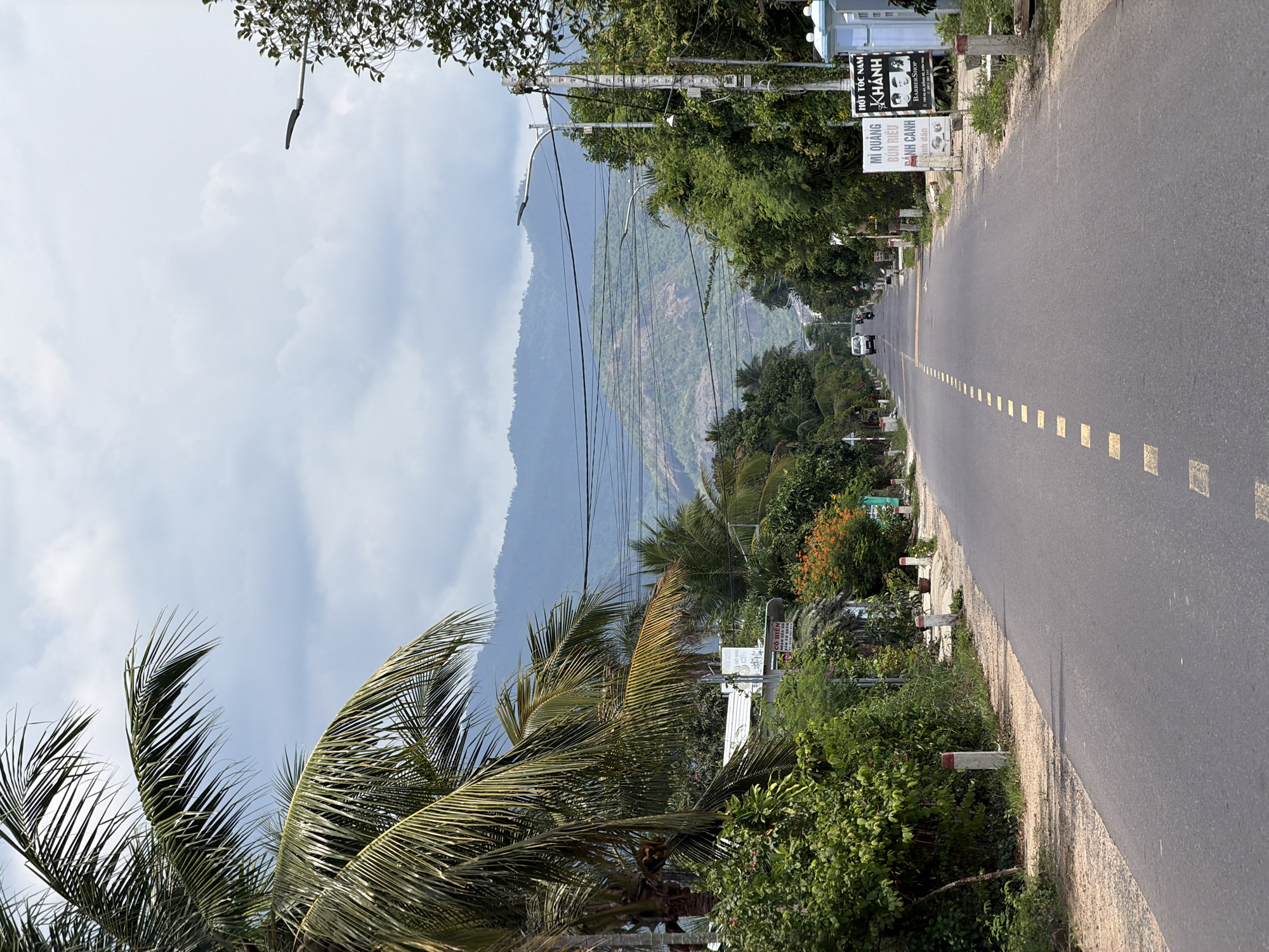 The image depicts a straight, asphalt road with a dashed yellow line down the center, extending into the distance. The road is flanked by lush green vegetation, palm trees, and various trees and bushes. Several concrete posts with red and white markings are visible along the roadside. On the right side of the road, there are signs, one of which advertises "MÌ QUẢNG BÚN RIÊU ĐANH CANH" and another, "HỚT TÓC NAM KHÁNH BARBERSHOP." In the background, a white car and several motorcycles are traveling on the road. The horizon is dominated by rolling, green mountains under a cloudy sky. The scene is set in Diên Thọ, Vietnam, likely a rural or semi-rural area, given the natural landscape and roadside businesses.