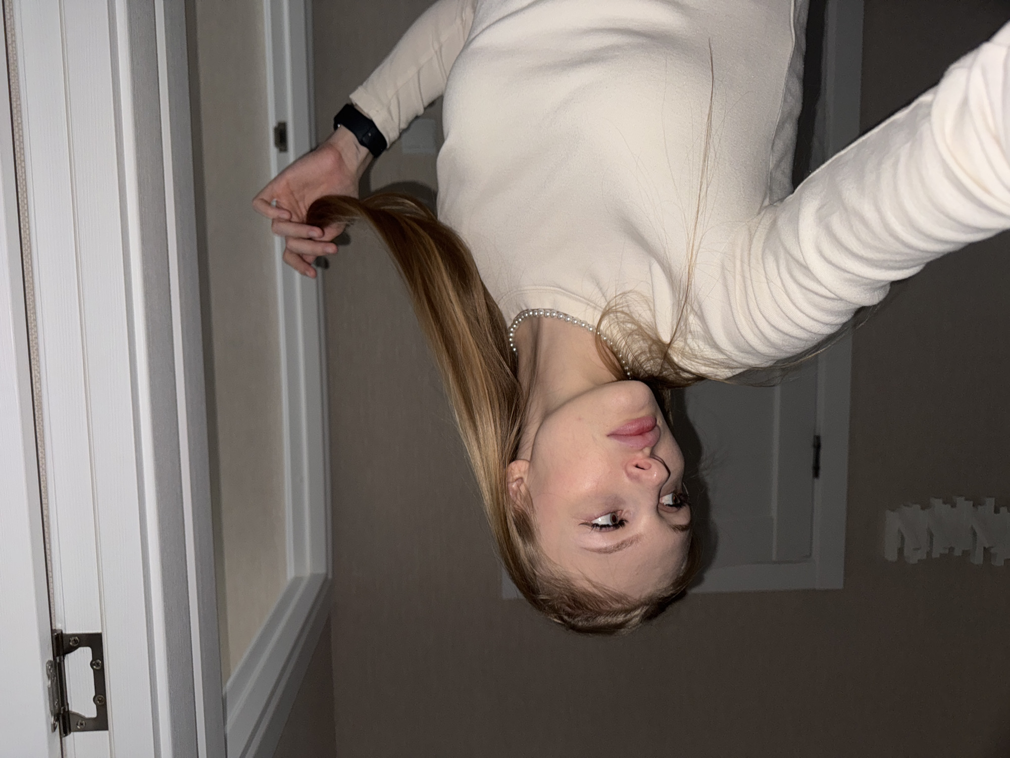 A young adult female is shown in a medium shot, indoors. She has blonde hair and is looking away from the camera to her left. She is wearing a cream-colored, long-sleeved shirt and a pearl necklace. Her right hand is holding a section of her hair. A black watch is visible on her left wrist. The background includes a doorway with white trim, and a wall with some decorative elements. The lighting suggests it is nighttime or a dimly lit room. There is no indication of the city of Perm, Russia in the image.