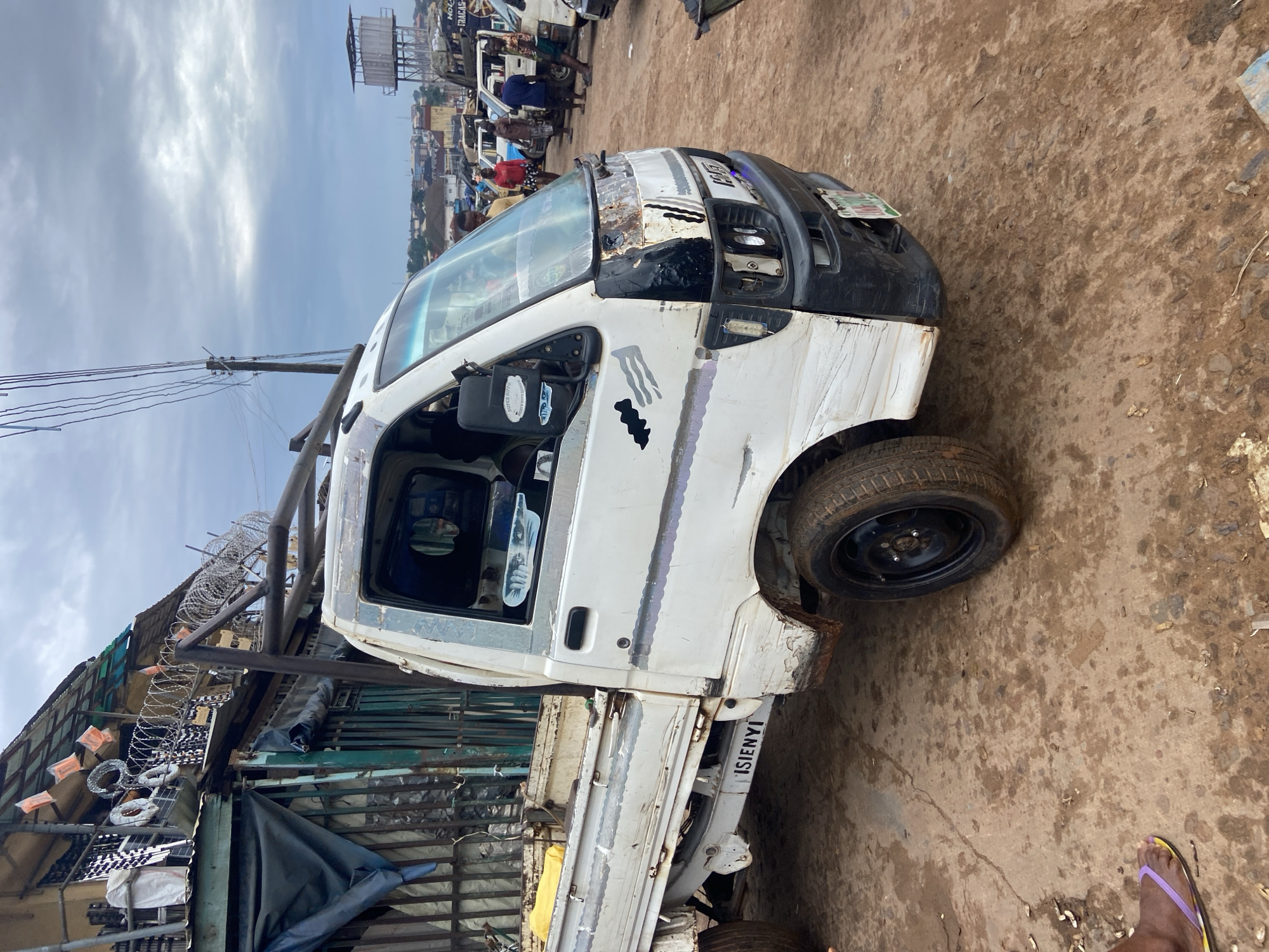 This media file shows a white, dilapidated truck parked on a dirt road in a market setting. The truck appears to be a utility vehicle, with a visible cargo bed and a driver's cabin. The vehicle has significant wear and tear, with rust and dents visible on its body. The front of the truck shows damage to the bumper and grille. A license plate is partially visible on the front.
In the background, other vehicles and market stalls are present, suggesting a busy commercial area. The sky is cloudy, indicating overcast weather. Power lines and poles are also visible, along with some barbed wire fencing and metal structures that may be part of the market.
Several people are present in the scene, though their activities are not clearly defined. One person is visible near the front of the truck, and others are in the background among the market stalls. A person's foot wearing a purple and yellow flip-flop is visible in the lower right corner of the image.
Visible text includes what appears to be writing on the side of the truck, possibly a name or identifier: "ISIENYI". Some stickers are also visible on the driver's side mirror, including one that reads "SUPRA AUTO". The overall impression is that of a functioning but worn-out vehicle in a bustling, informal market environment.