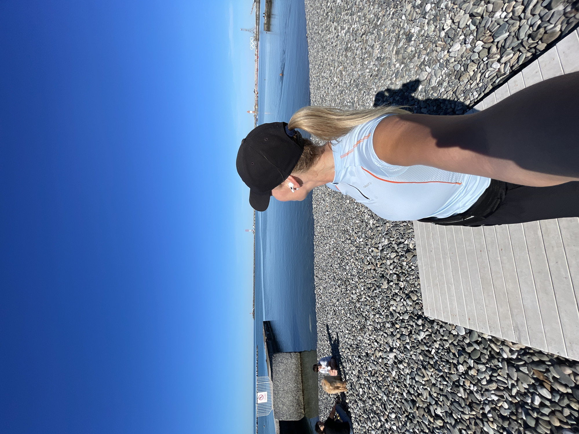 The image, taken in Sochi, Russia, captures a bright, sunny day at a pebbly beach alongside calm blue water. The primary subject is a person, likely female, viewed from the waist up in profile, facing left towards the sea. She wears a black baseball cap, a light blue short-sleeved athletic shirt with orange accents (a "CRAFT" logo is visible), and white wireless earphones. Her blonde hair is pulled back into a ponytail that extends from under her cap.
The setting features a wide expanse of clear blue sky dominating the upper left, meeting the horizon where the sea begins. The water is a deep blue, reflecting the sky. On the right side of the image, a light-colored wooden platform or walkway is visible, with a border of small, smooth grey and brown pebbles. Strong sunlight casts clear shadows from the person and other unseen objects onto the pebbled ground.
In the distance across the water, various structures can be seen, including what appears to be the iconic Olympic rings, a lighthouse, and other buildings, hinting at an urban or developed coastline. Further down the beach (visible in wider crops), other individuals and a dog are present. A sign near the water, though partially obscured, appears to show a "NO DIVING" symbol with a crossed-out swimmer. The overall atmosphere is serene and bright, characteristic of a pleasant day by the sea.