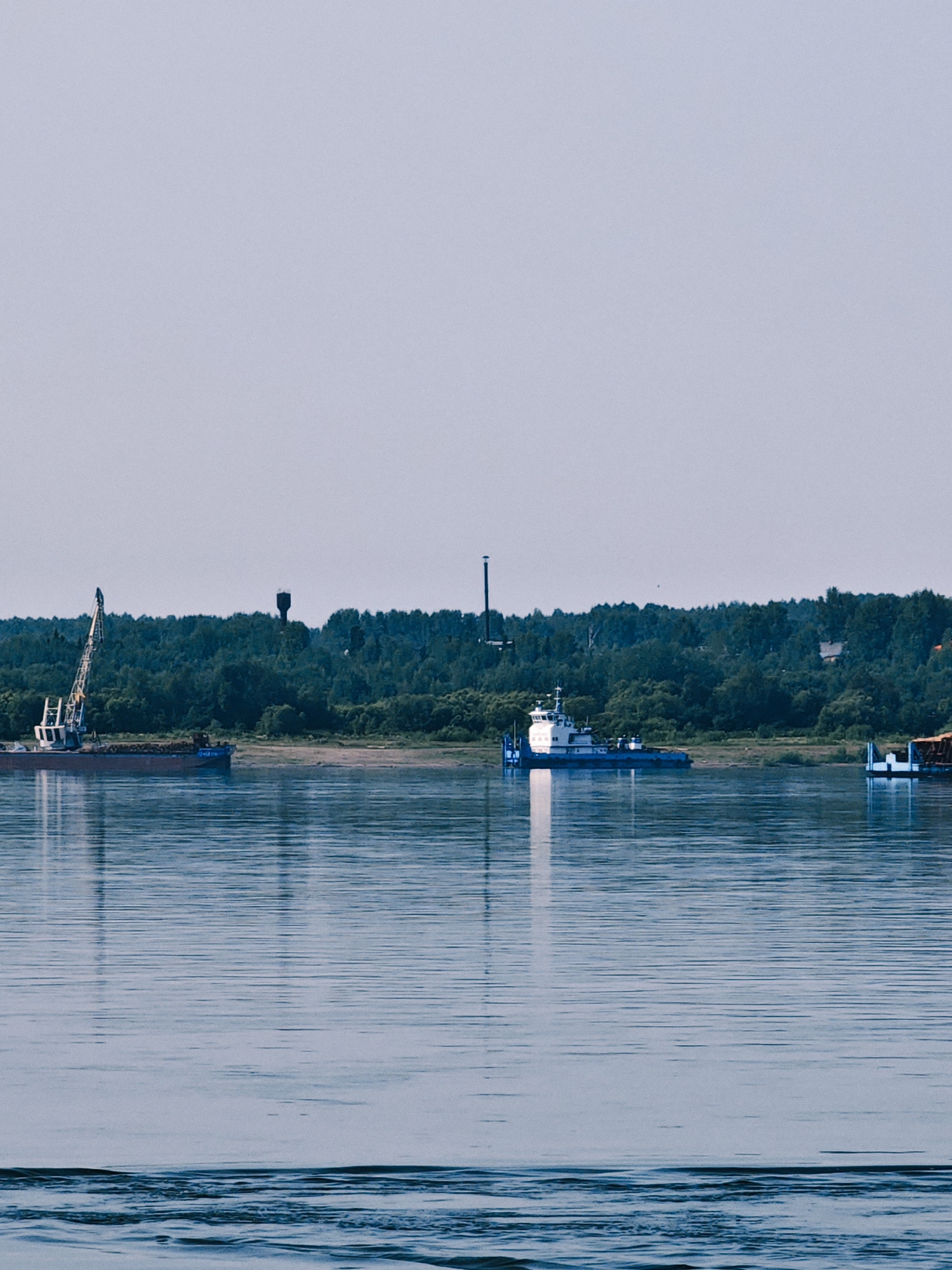 A wide, calm river flows in the foreground of the image. In the middle ground, a forested shoreline is visible, with trees of varying shades of green forming a dense canopy. A tall, dark water tower stands out against the trees. Several vessels are present on the river. On the left, a barge with a large crane is docked near the shore. Further to the right, a white and blue boat is navigating the water. On the far right, part of another barge, possibly carrying cargo, is visible. The sky above is a pale, hazy blue, suggesting it is daytime. The overall scene depicts a serene river landscape with industrial elements.