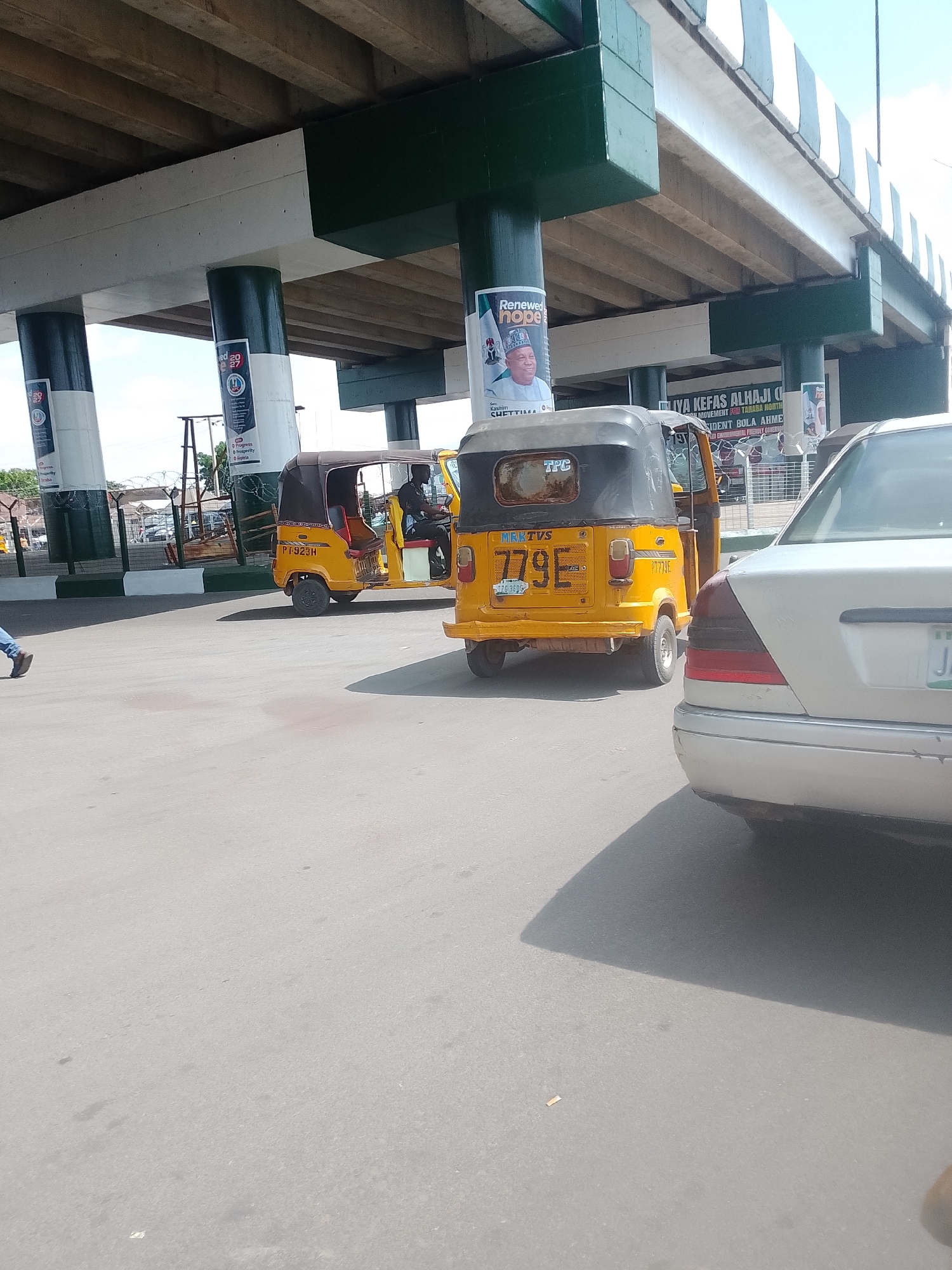 This is a street-level, outdoor, daytime photograph taken in Jalingo, Nigeria. The image features a brightly colored yellow auto-rickshaw, commonly known as a "Keke," in the foreground, facing right. The auto-rickshaw has the license plate "PJ 923H" visible on its rear. To the right of the auto-rickshaw, a silver sedan is partially visible, its rear quarter panel and taillight in focus. In the background, a large concrete overpass supported by dark green and white pillars dominates the scene. Several posters with political campaign messaging are affixed to the pillars. The ground is paved, and the overall impression is one of urban street activity.